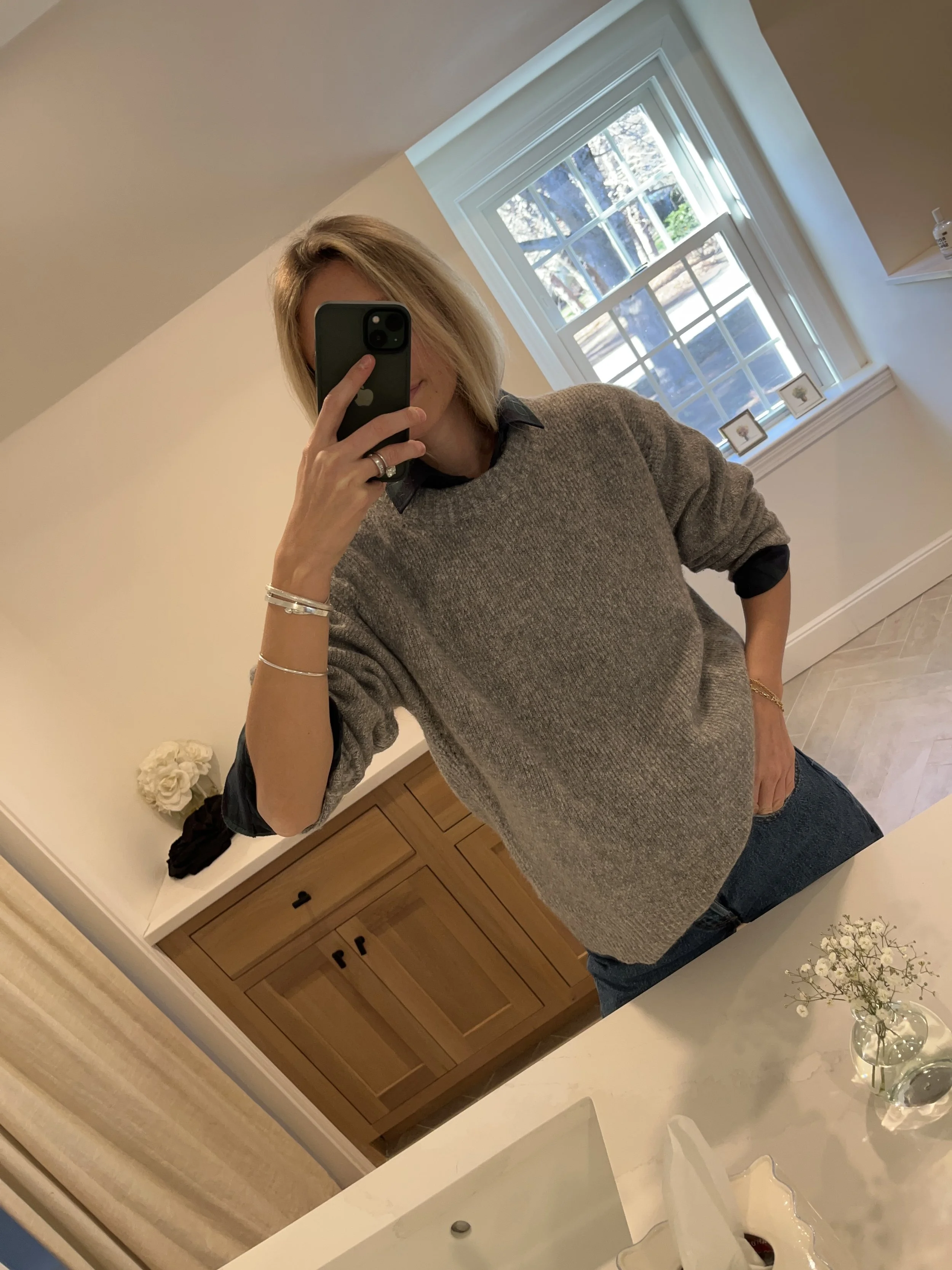 A woman taking a mirror selfie in a spacious living room with wooden floors, large windows, and minimal furniture, wearing a beige jacket, black top, blue jeans, and tan shoes.