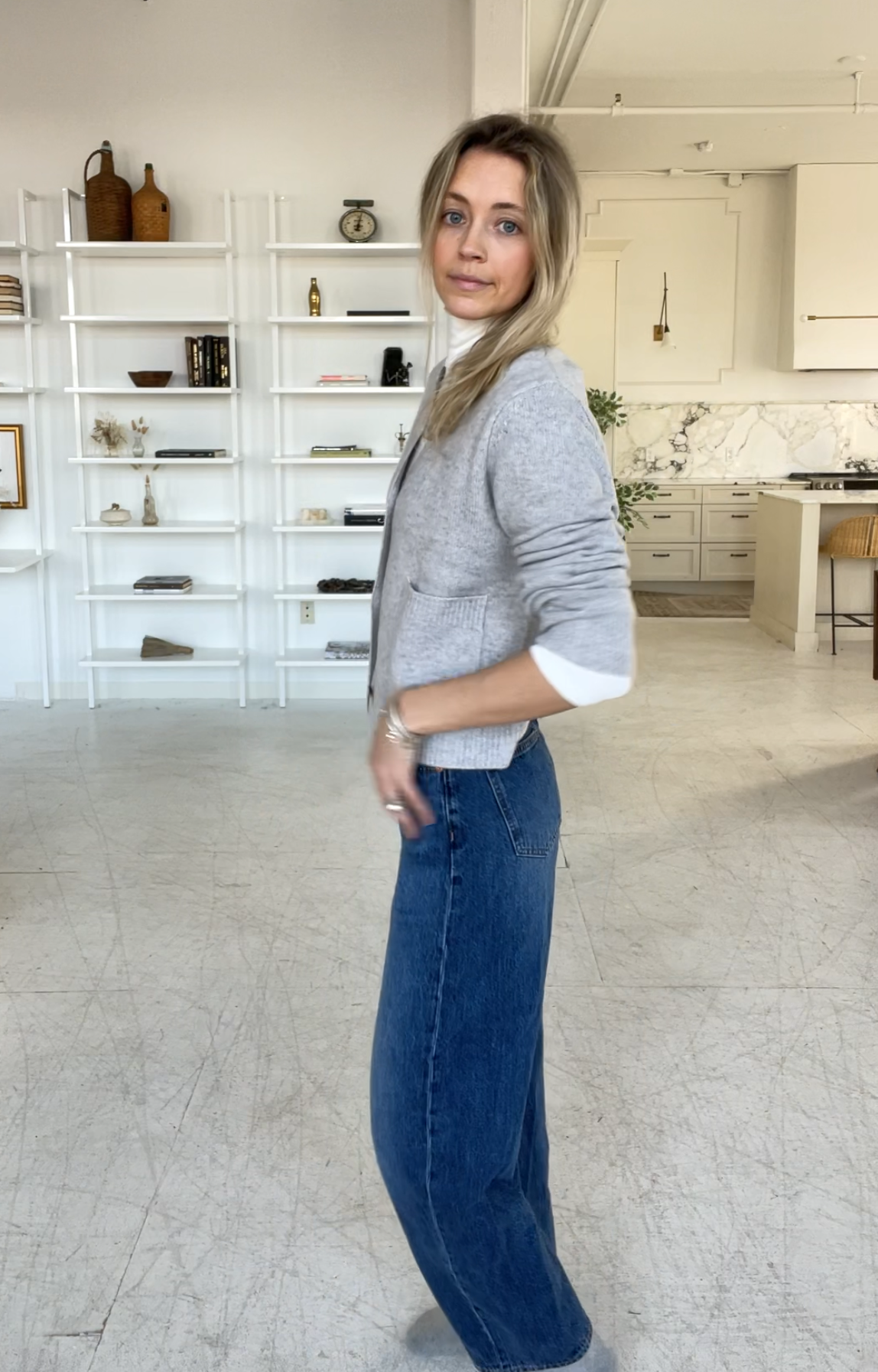 Person taking a mirror selfie in a room with wooden floors, wearing a striped brown and white long-sleeve shirt, light blue jeans, and black and white sneakers.