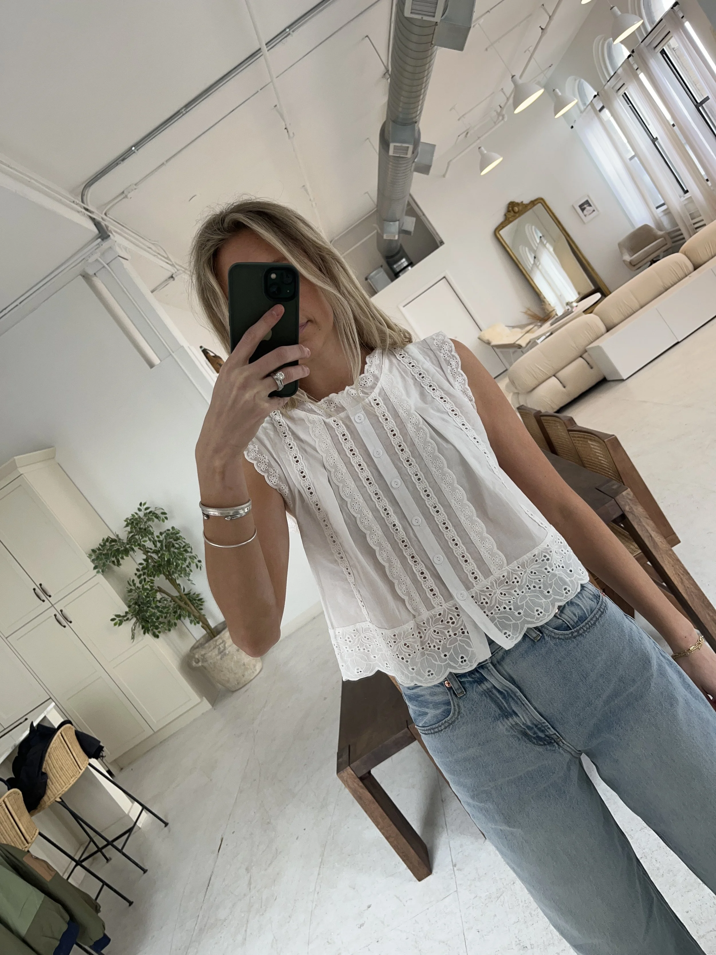 A woman taking a mirror selfie in a spacious living room with wooden floors, large windows, and minimal furniture, wearing a beige jacket, black top, blue jeans, and tan shoes.