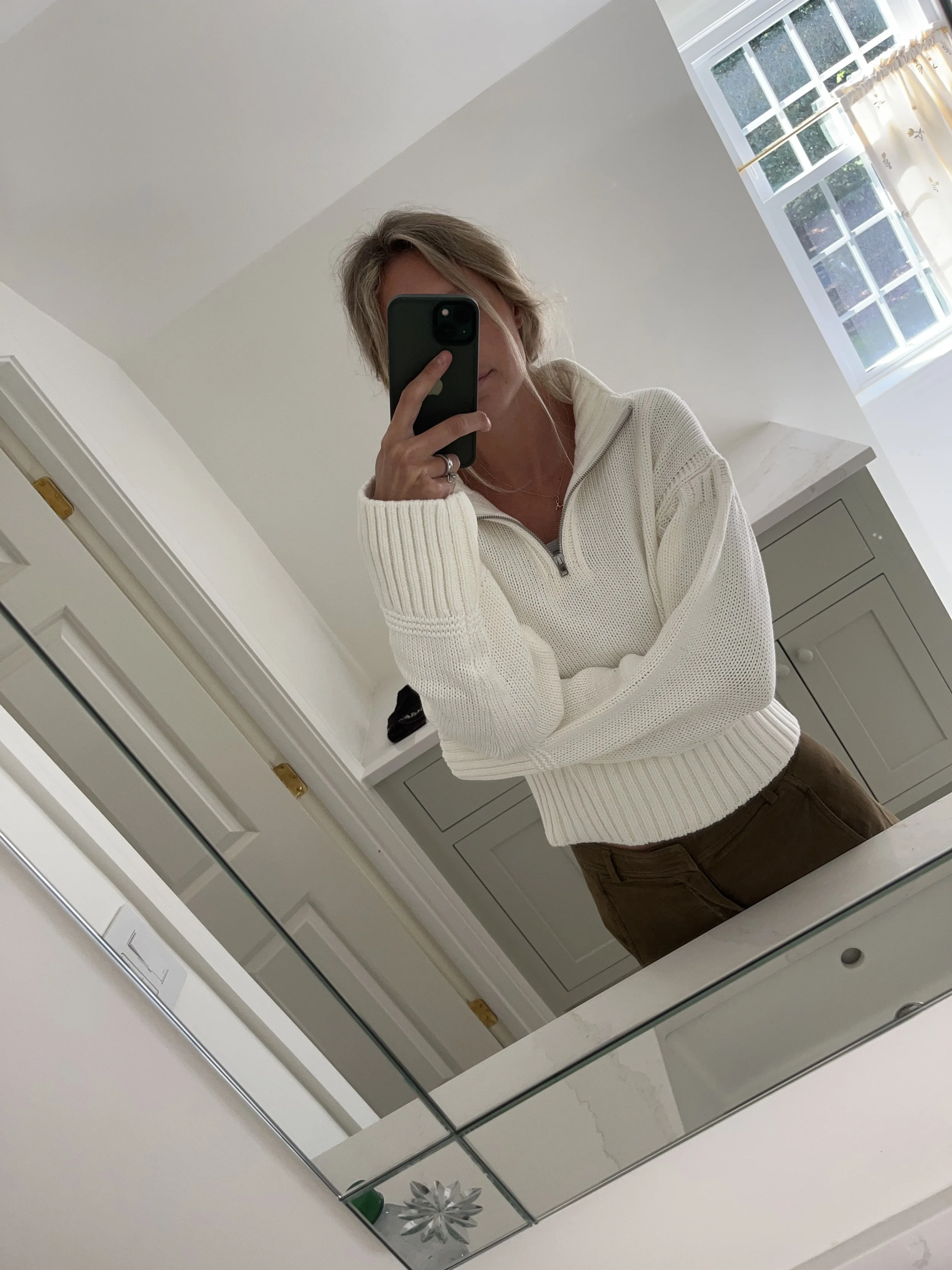 Person taking a mirror selfie in a living room with hardwood floors, wearing a black fleece jacket, gray shirt, black leggings, gray sneakers, and a white cap with a logo.