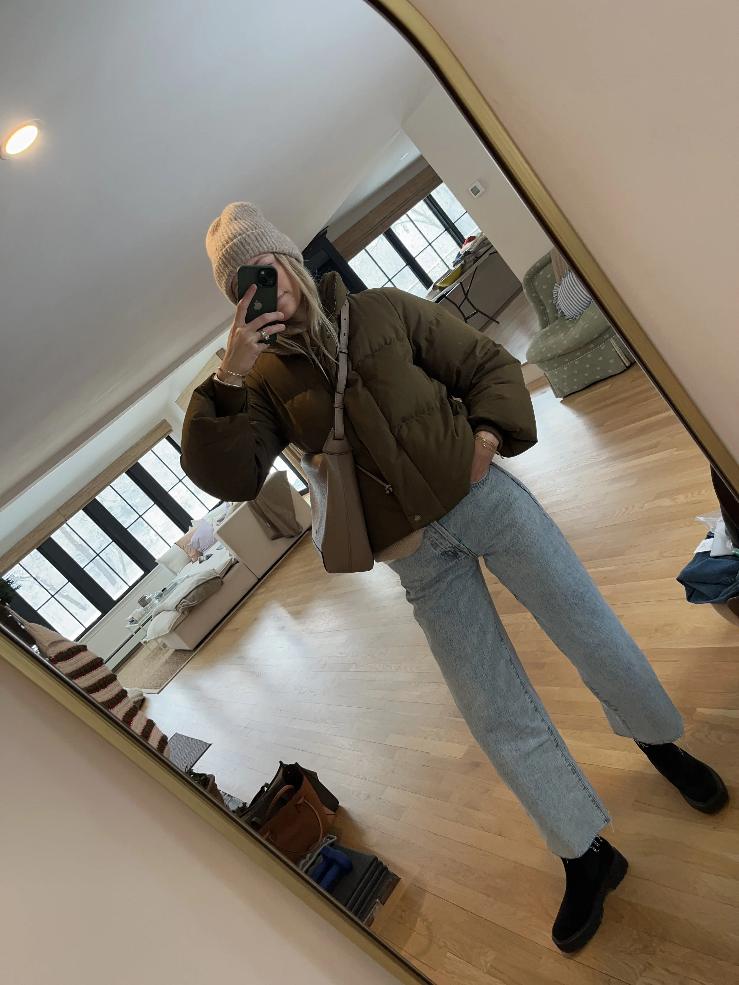 Person taking a mirror selfie in a room with wooden floors, wearing a striped brown and white long-sleeve shirt, light blue jeans, and black and white sneakers.