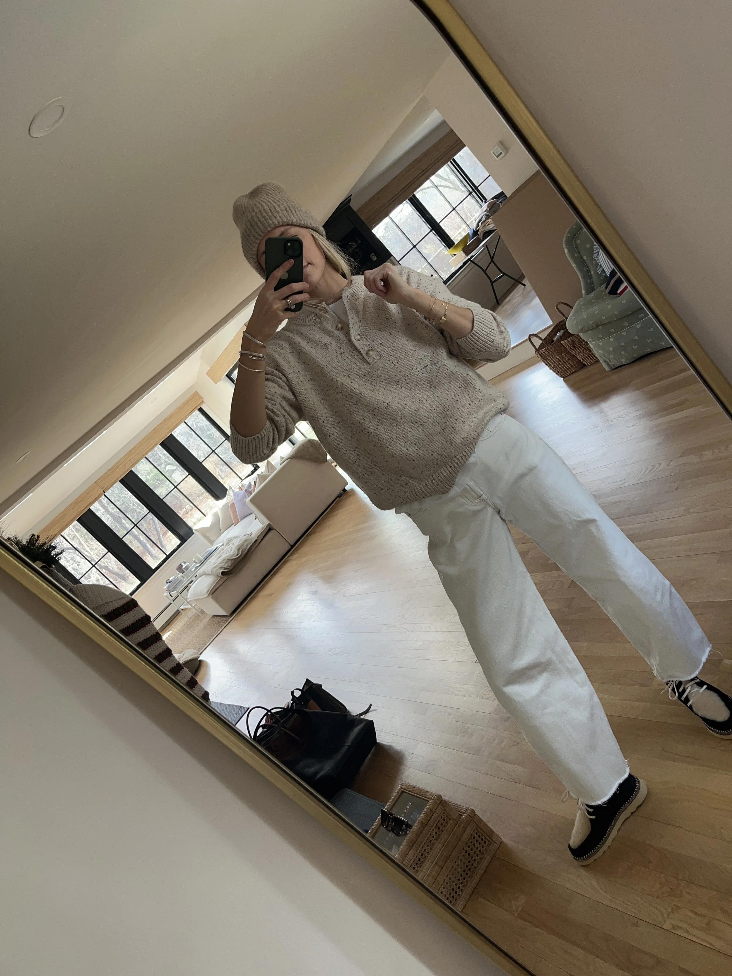 Person taking a mirror selfie in a room with wooden floors, wearing a striped brown and white long-sleeve shirt, light blue jeans, and black and white sneakers.