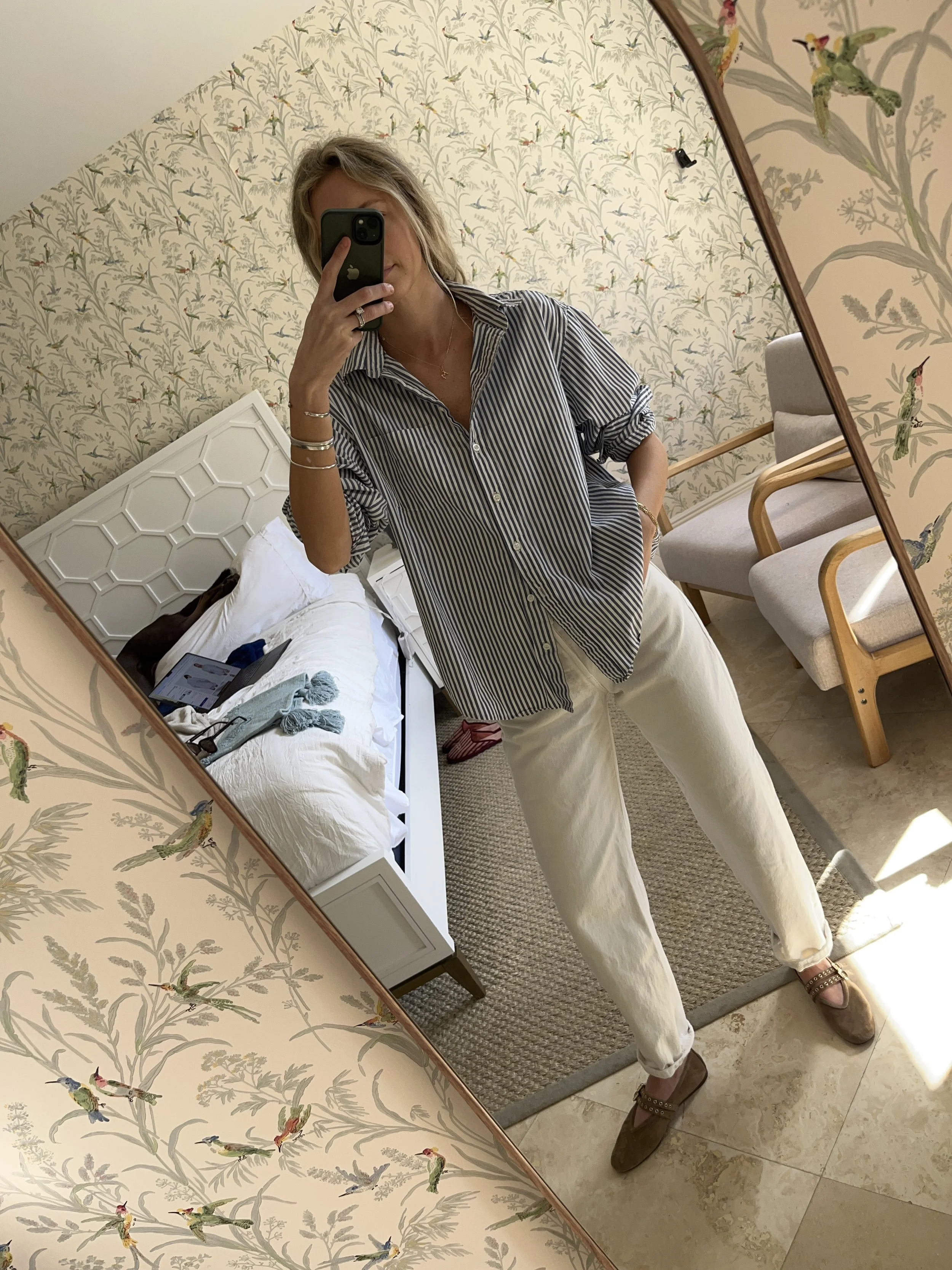A woman taking a mirror selfie in a spacious living room with wooden floors, large windows, and minimal furniture, wearing a beige jacket, black top, blue jeans, and tan shoes.