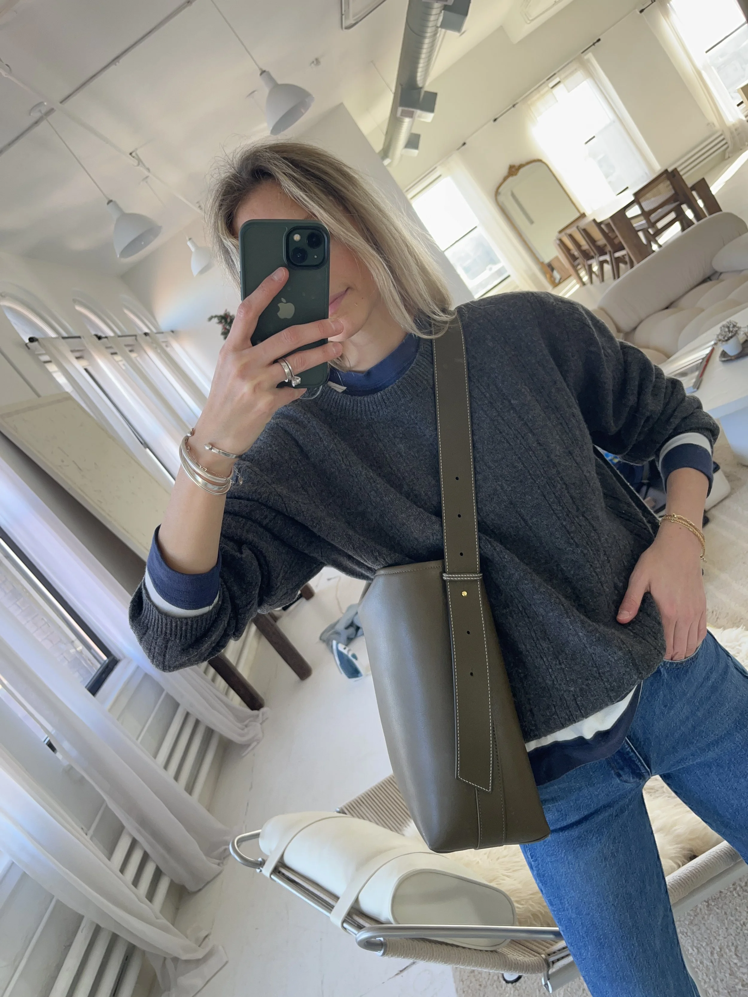 Person taking a mirror selfie in a room with wooden floors, wearing a striped brown and white long-sleeve shirt, light blue jeans, and black and white sneakers.