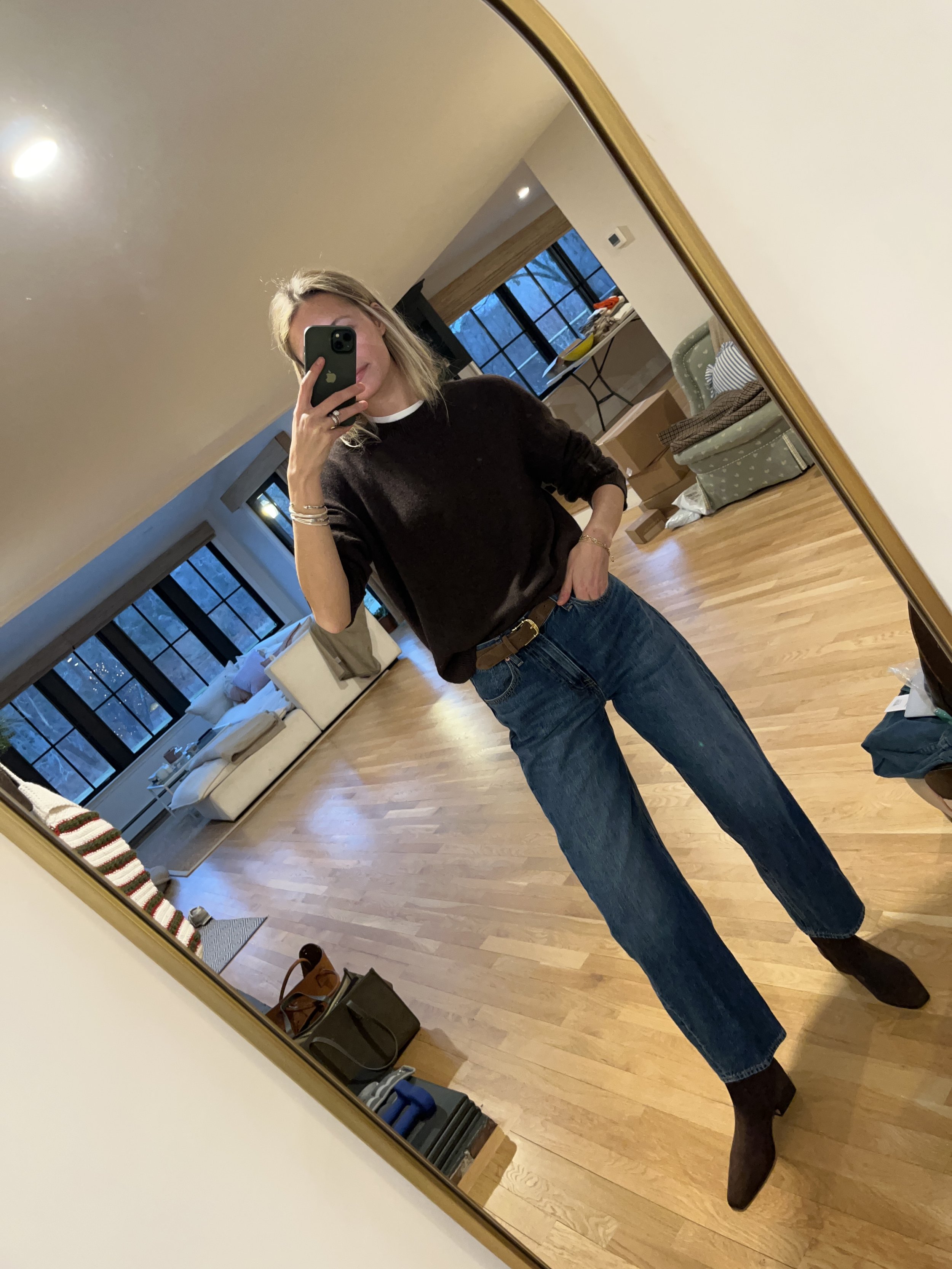 Person taking a mirror selfie in a living room with hardwood floors, wearing a black fleece jacket, gray shirt, black leggings, gray sneakers, and a white cap with a logo.