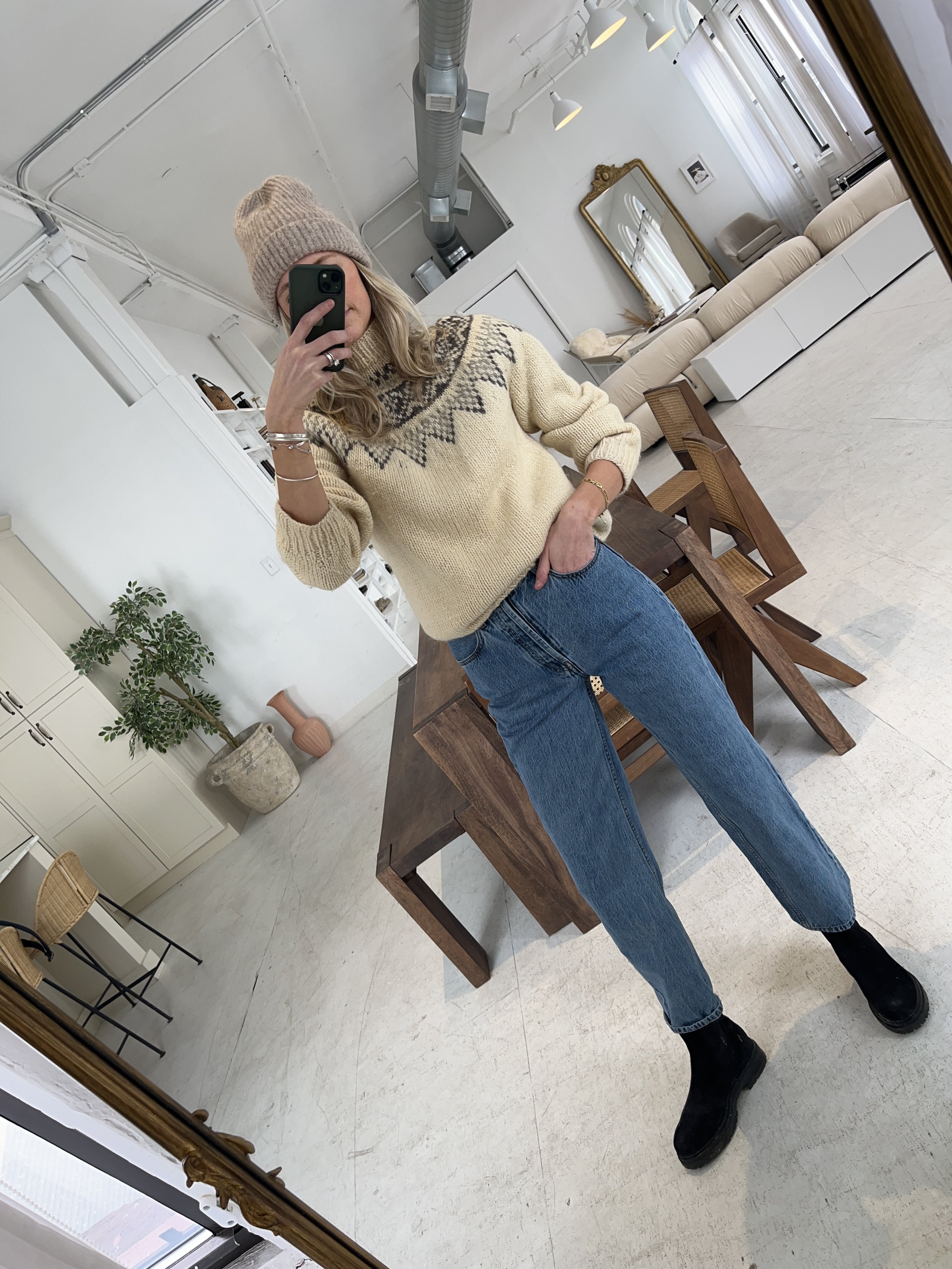 Person taking a mirror selfie in a living room with hardwood floors, wearing a black fleece jacket, gray shirt, black leggings, gray sneakers, and a white cap with a logo.