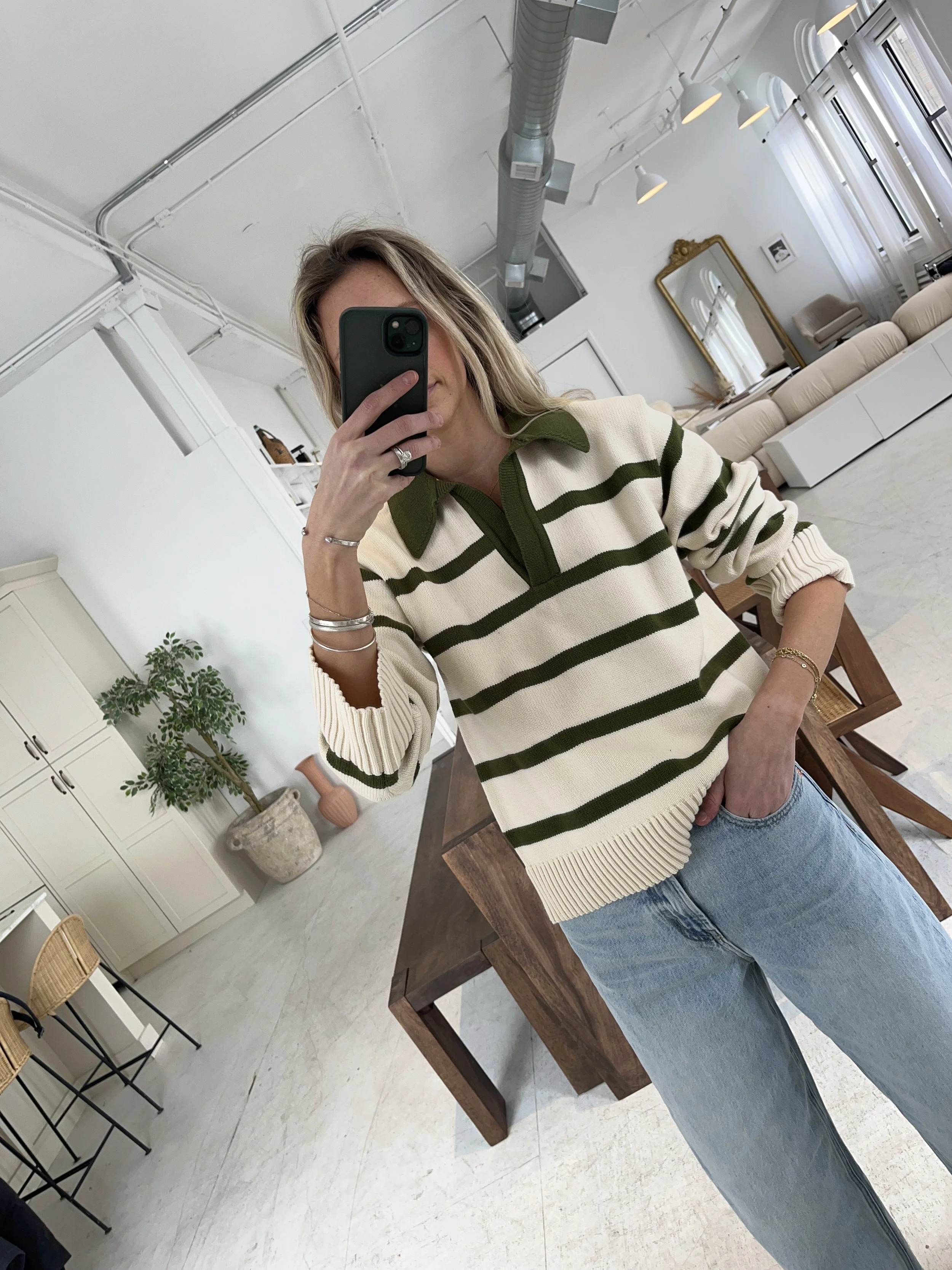 A woman taking a mirror selfie in a spacious, well-lit living room with hardwood floors, large windows, a white sofa, and a side table with a plant.