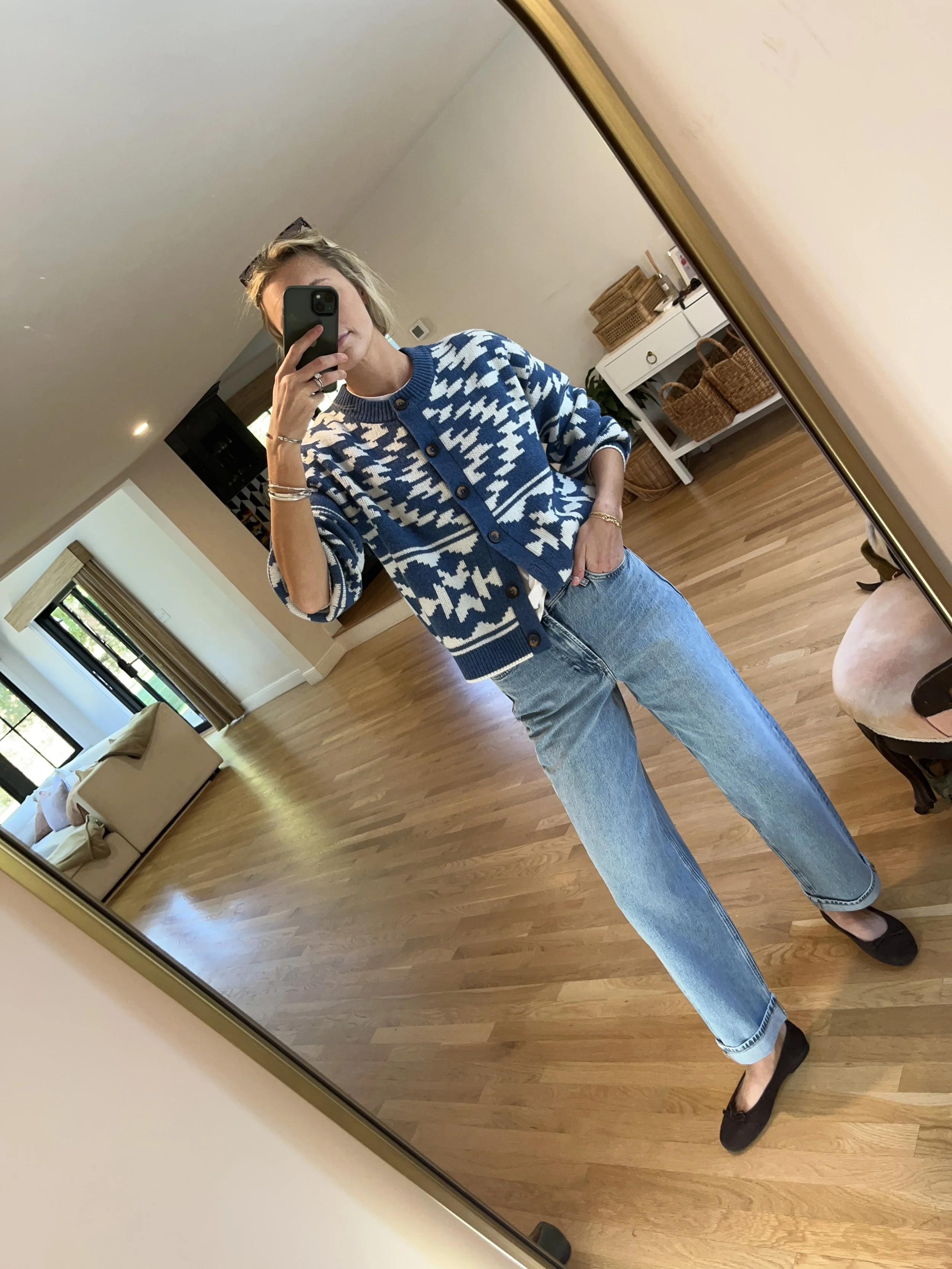 A woman taking a mirror selfie in a spacious living room with wooden floors, large windows, and minimal furniture, wearing a beige jacket, black top, blue jeans, and tan shoes.