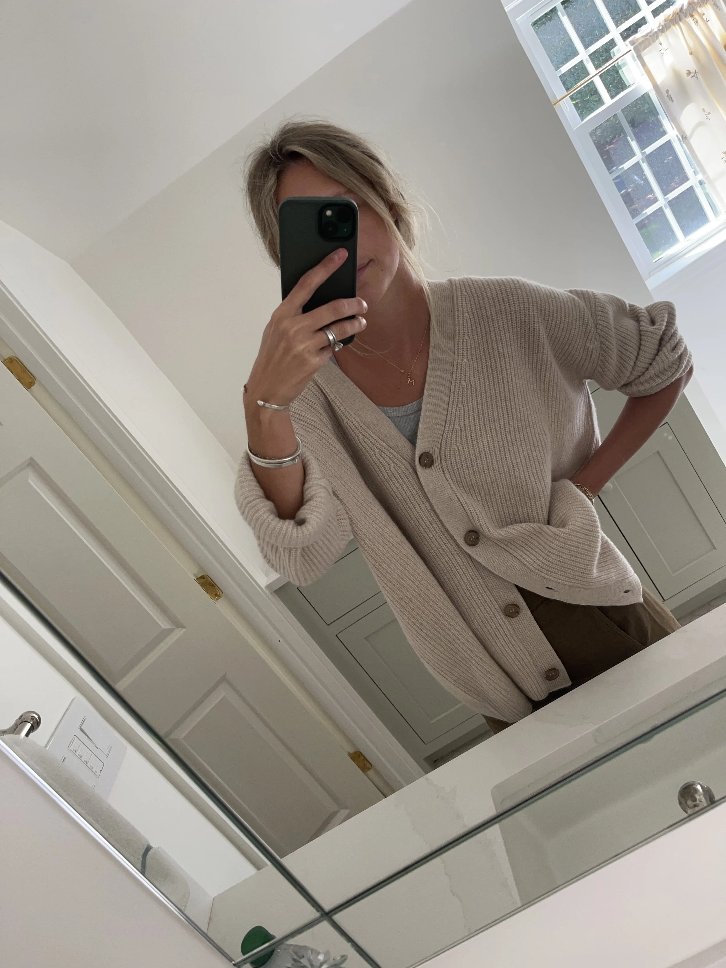 Person taking a mirror selfie in a room with wooden floors, wearing a striped brown and white long-sleeve shirt, light blue jeans, and black and white sneakers.