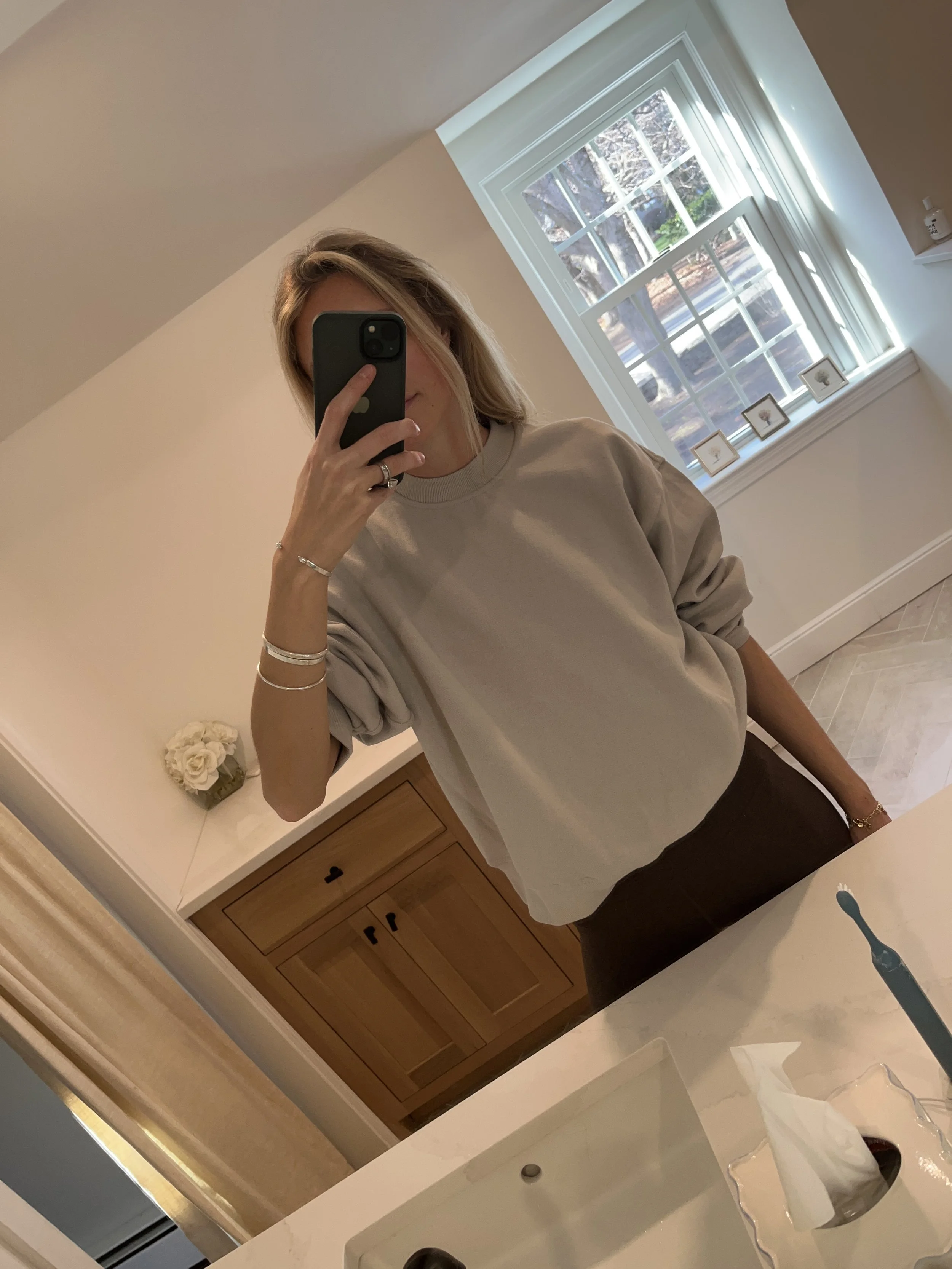 A woman taking a mirror selfie in a spacious, well-lit living room with hardwood floors, large windows, a white sofa, and a side table with a plant.