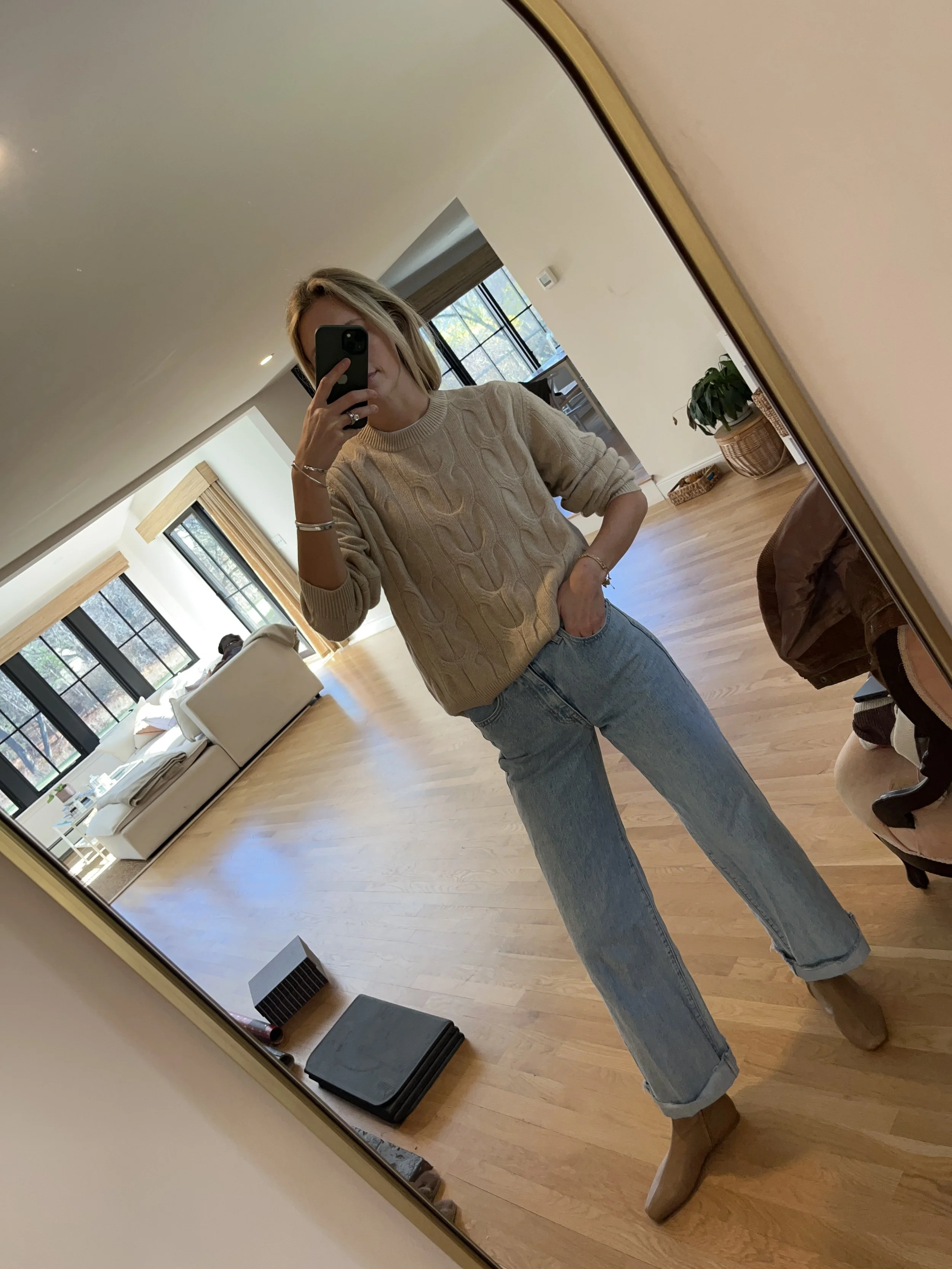A woman taking a mirror selfie in a spacious living room with wooden floors, large windows, and minimal furniture, wearing a beige jacket, black top, blue jeans, and tan shoes.