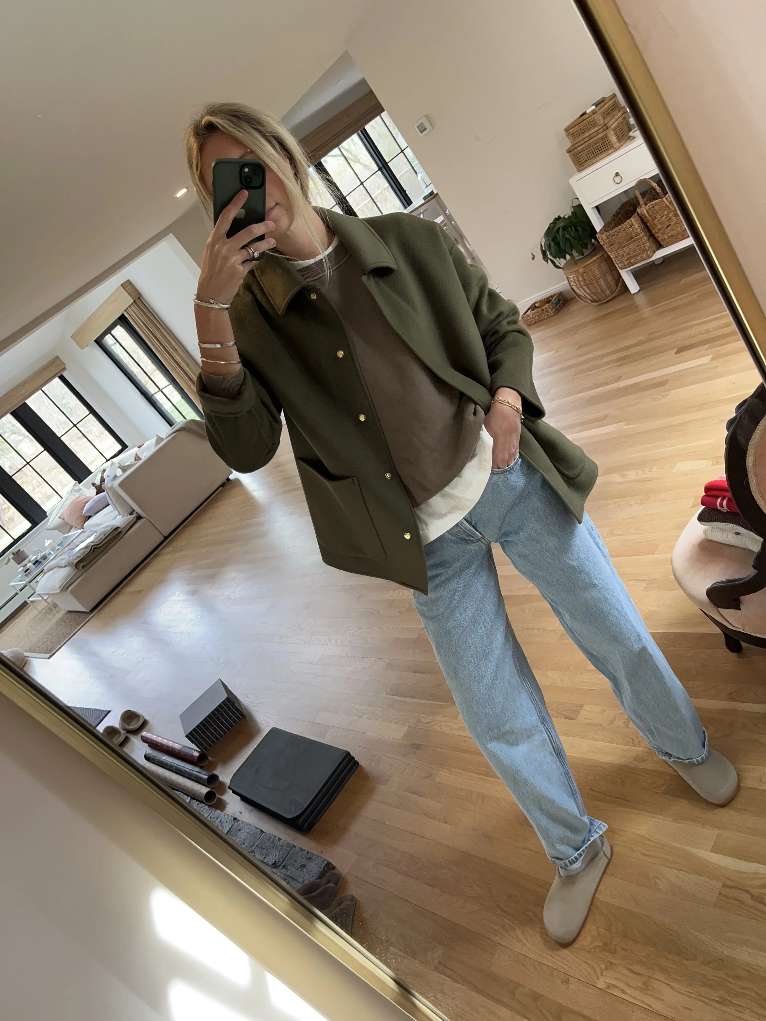 A woman taking a mirror selfie in a spacious, well-lit living room with hardwood floors, large windows, a white sofa, and a side table with a plant.