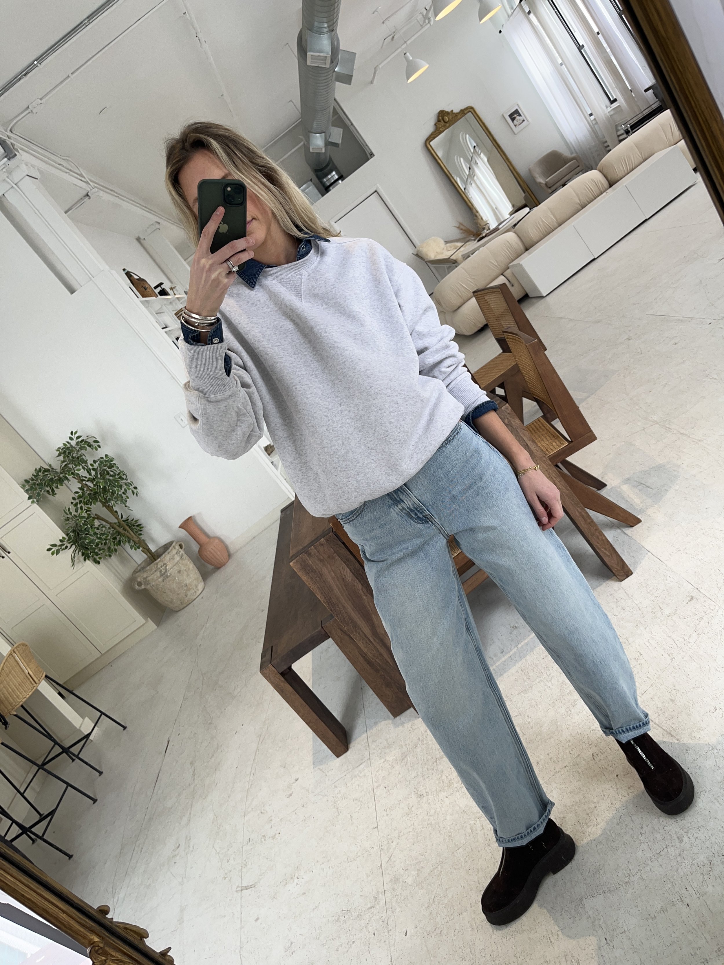 A woman taking a mirror selfie in a spacious living room with wooden floors, large windows, and minimal furniture, wearing a beige jacket, black top, blue jeans, and tan shoes.