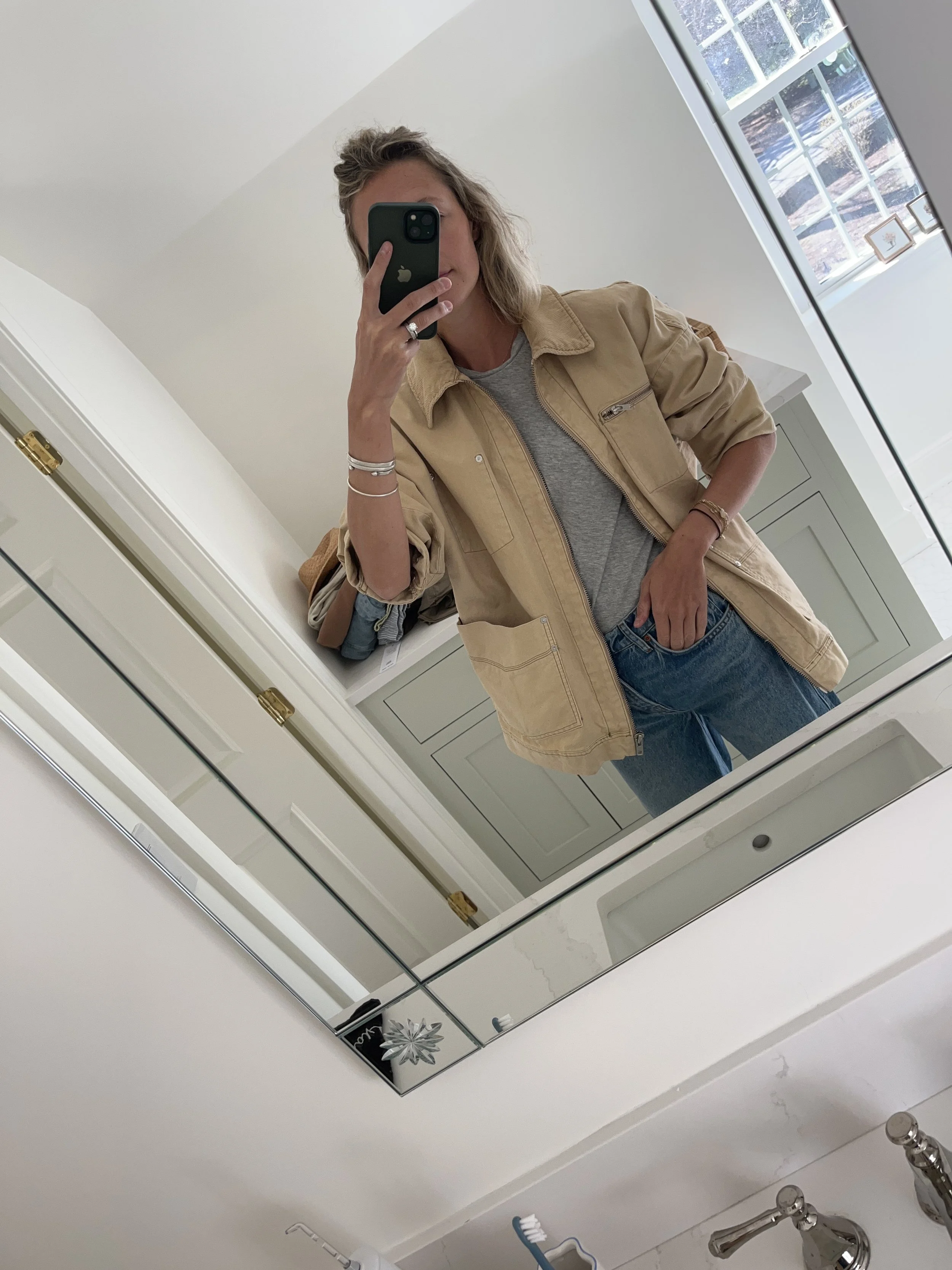 A woman taking a mirror selfie in a spacious living room with wooden floors, large windows, and minimal furniture, wearing a beige jacket, black top, blue jeans, and tan shoes.