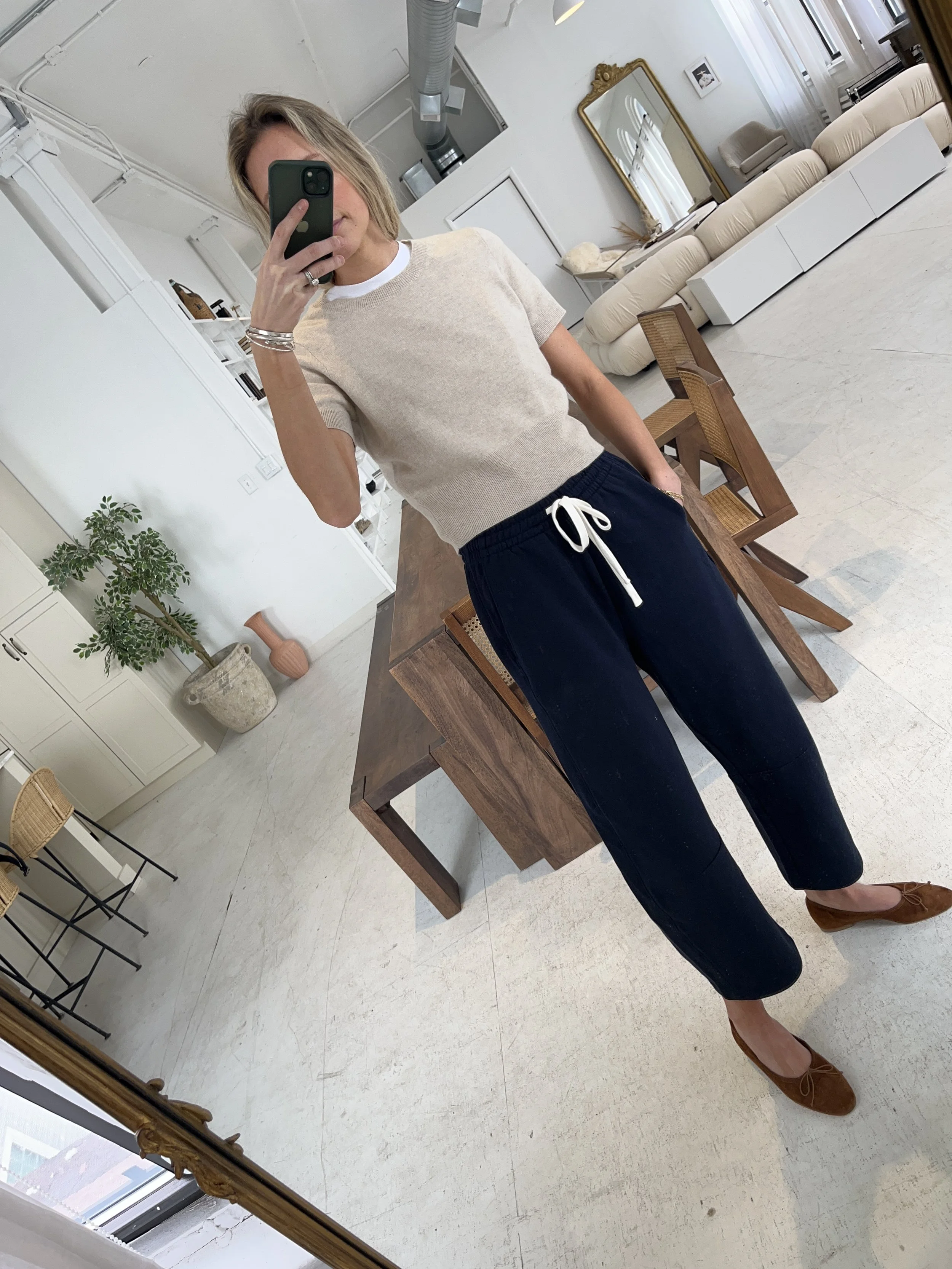 Person taking a mirror selfie in a room with wooden floors, wearing a striped brown and white long-sleeve shirt, light blue jeans, and black and white sneakers.