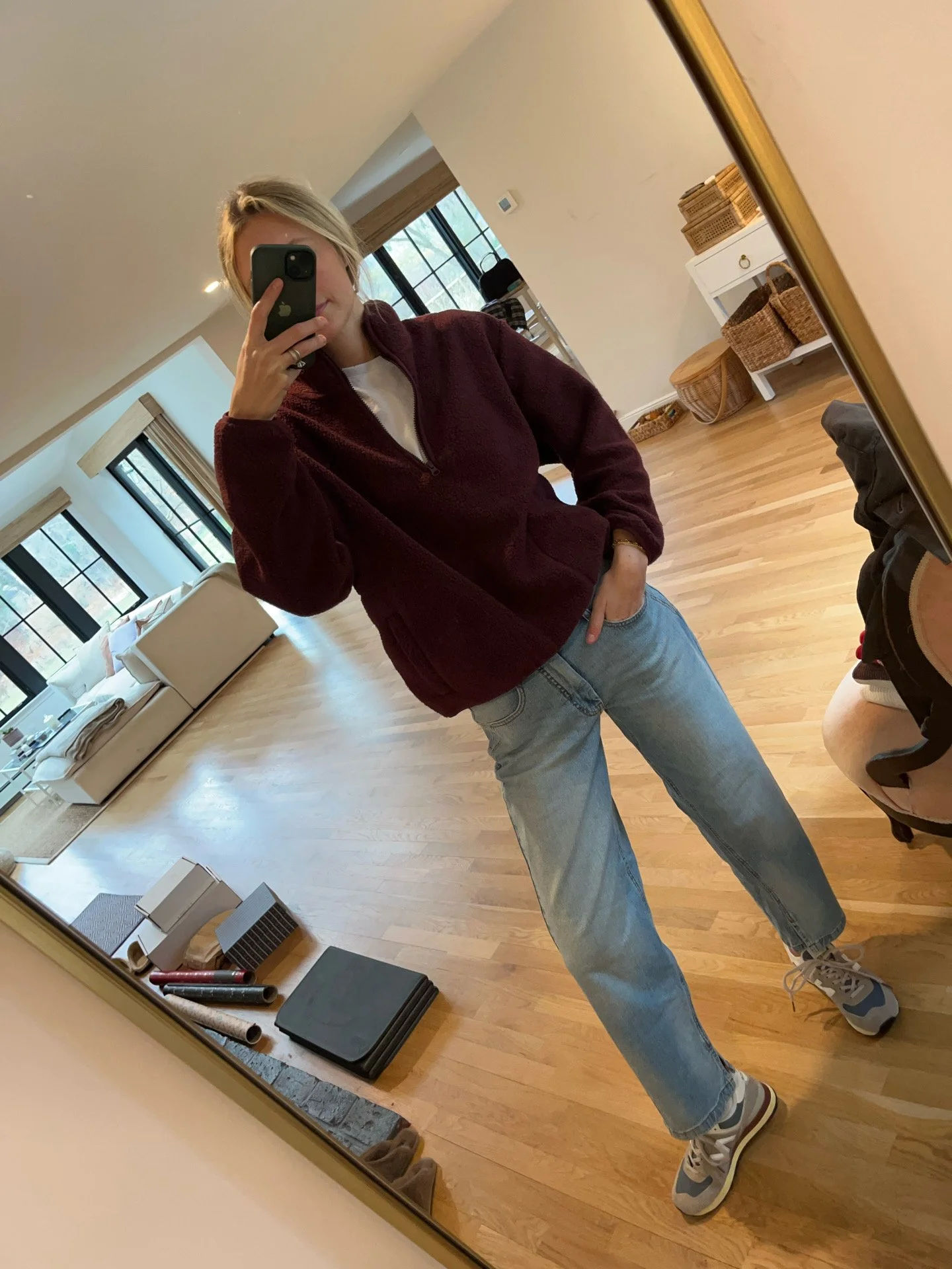 Person taking a mirror selfie in a living room with hardwood floors, wearing a black fleece jacket, gray shirt, black leggings, gray sneakers, and a white cap with a logo.