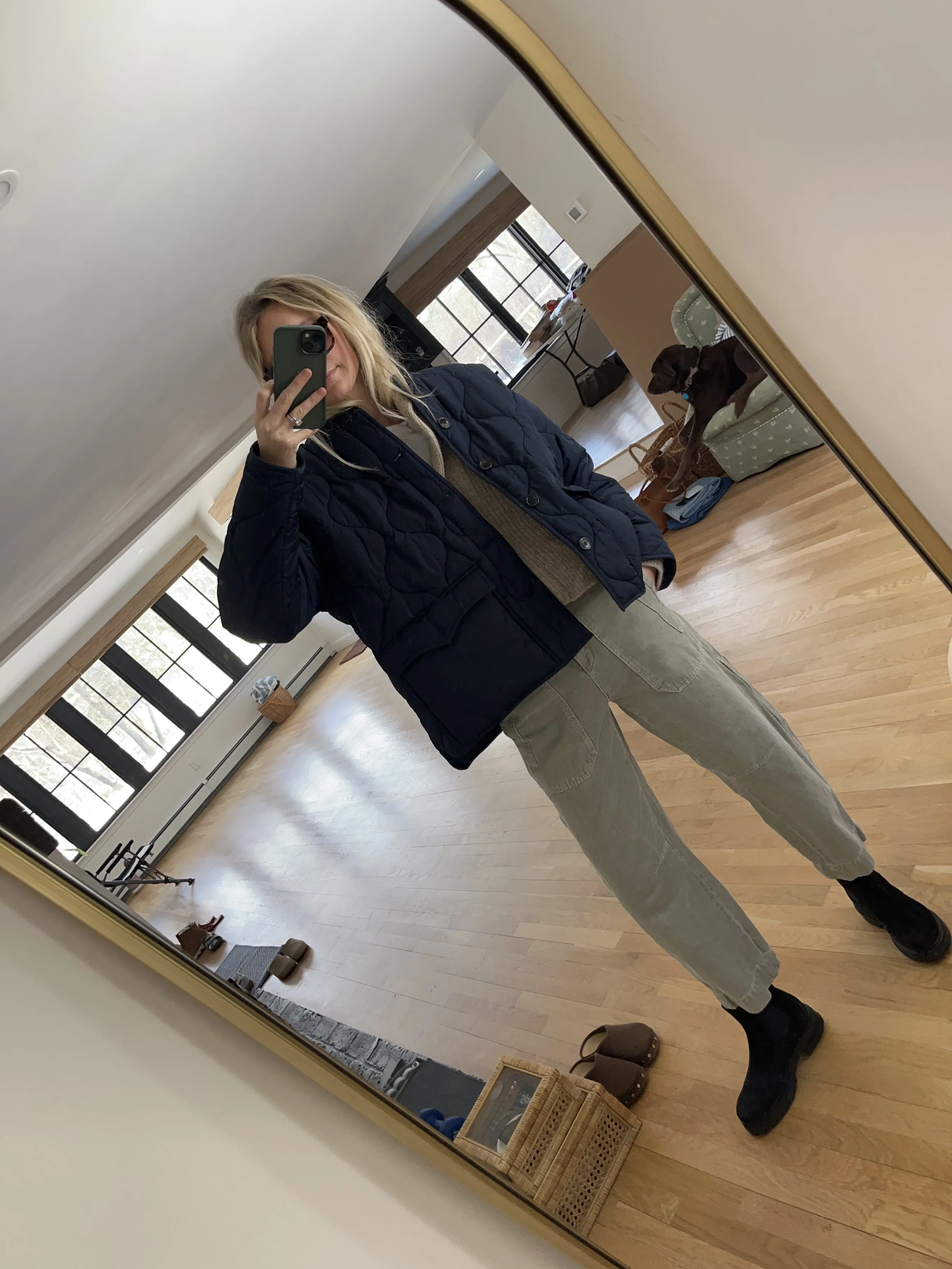 Person taking a mirror selfie in a room with wooden floors, wearing a striped brown and white long-sleeve shirt, light blue jeans, and black and white sneakers.