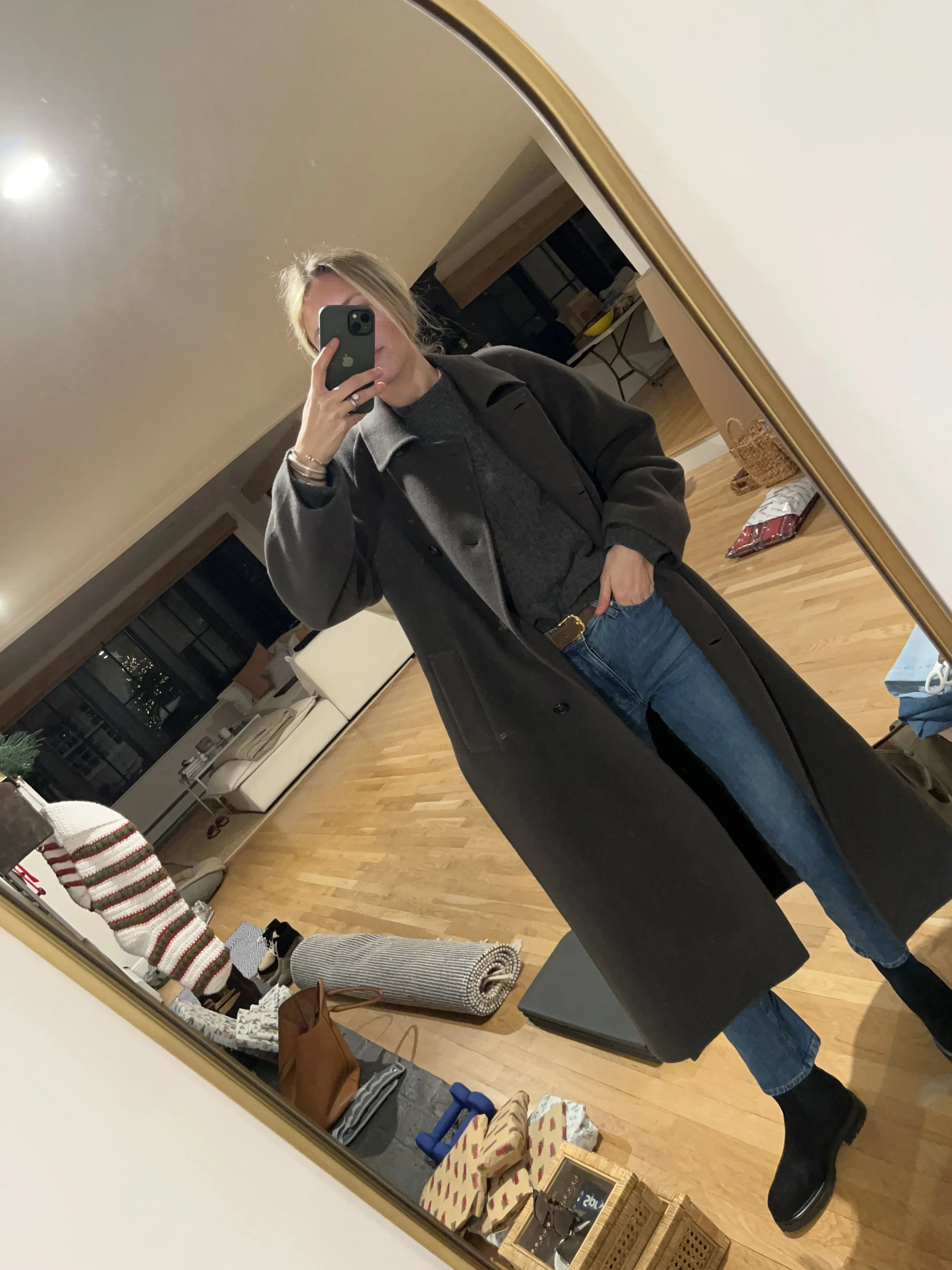 A woman taking a mirror selfie in a spacious, well-lit living room with hardwood floors, large windows, a white sofa, and a side table with a plant.