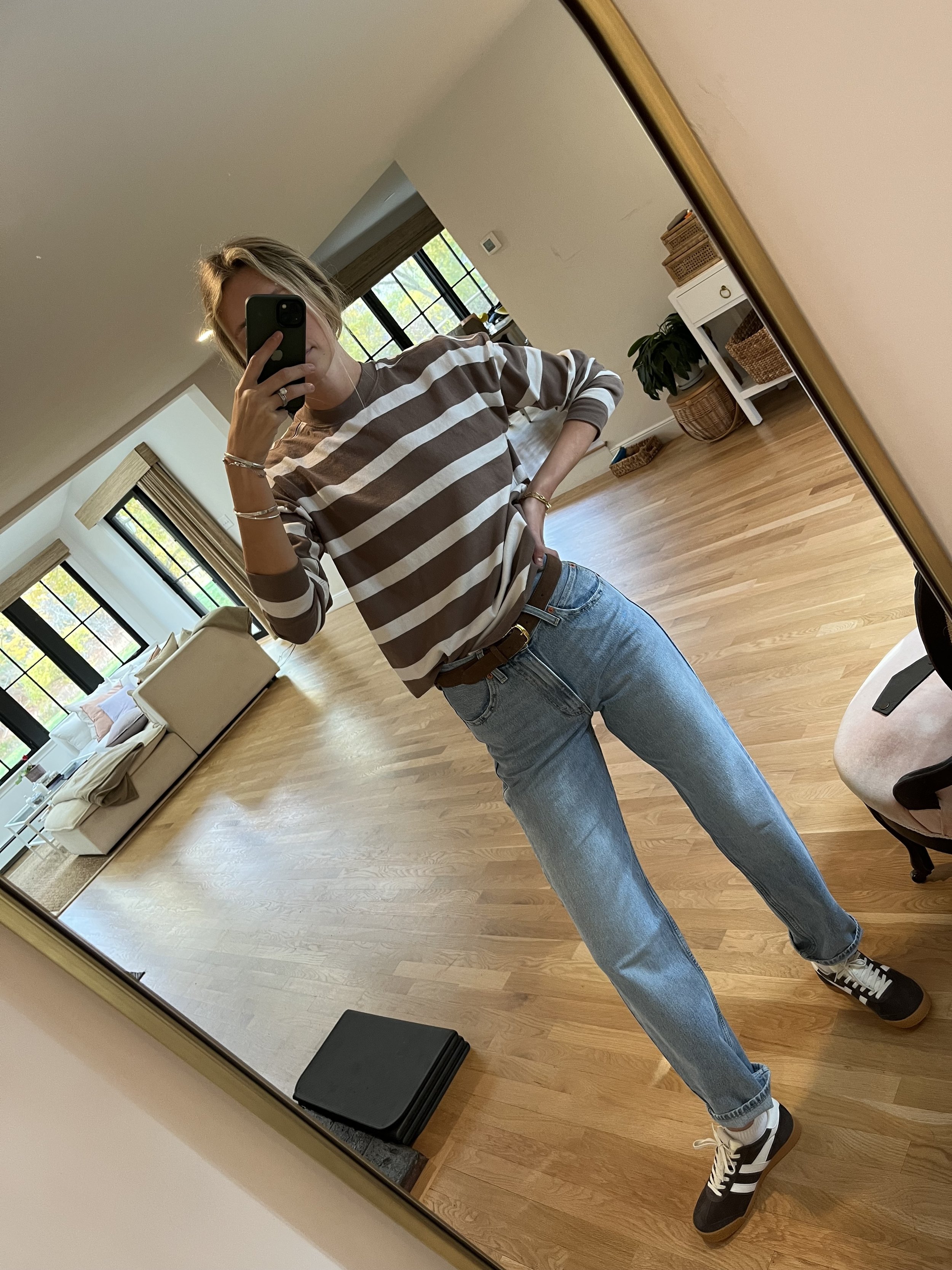 Person taking a mirror selfie in a room with wooden floors, wearing a striped brown and white long-sleeve shirt, light blue jeans, and black and white sneakers.