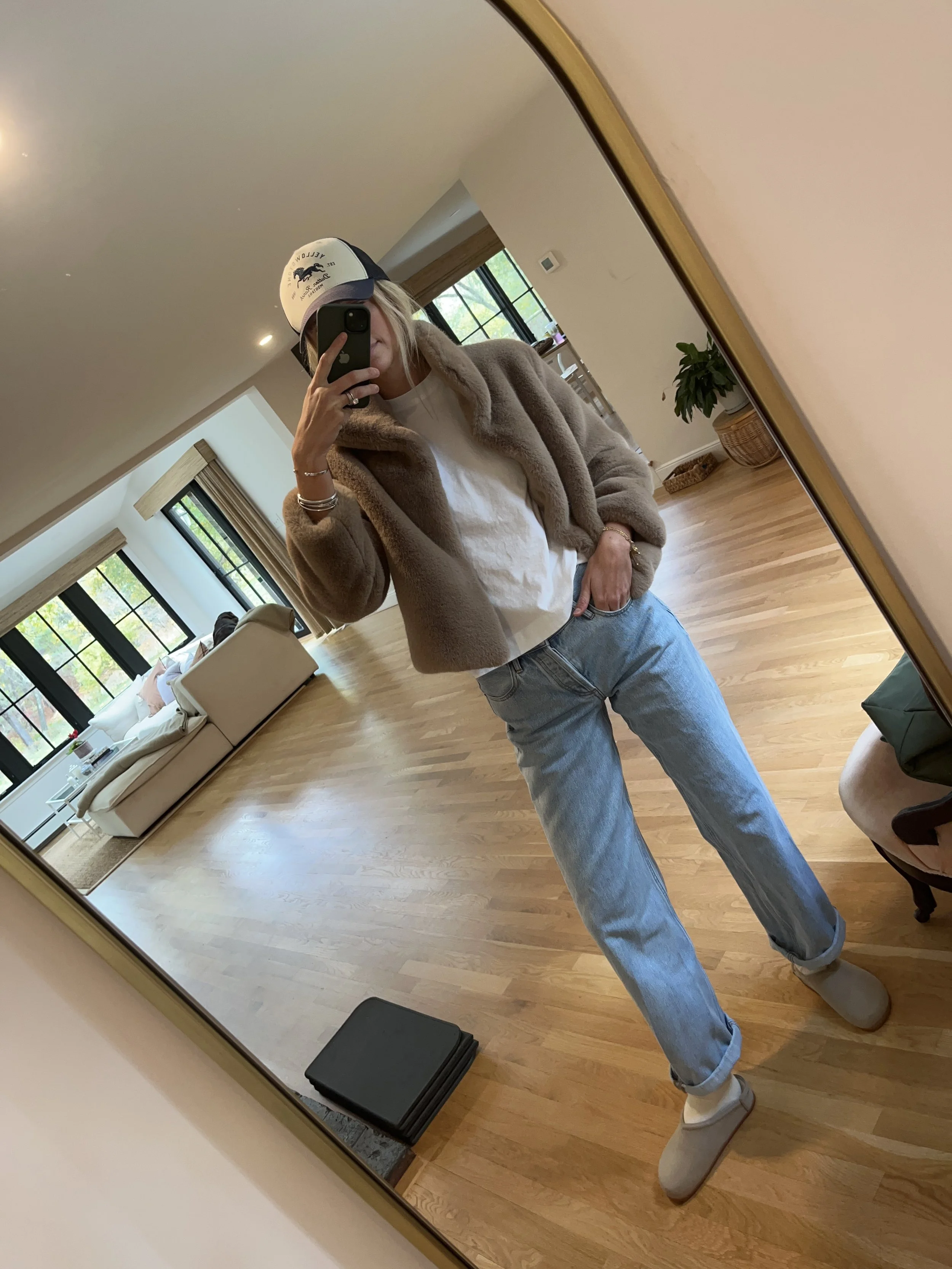 Person taking a mirror selfie in a spacious living room, wearing a beige fur jacket, white shirt, light blue jeans, beige shoes, and a white cap.