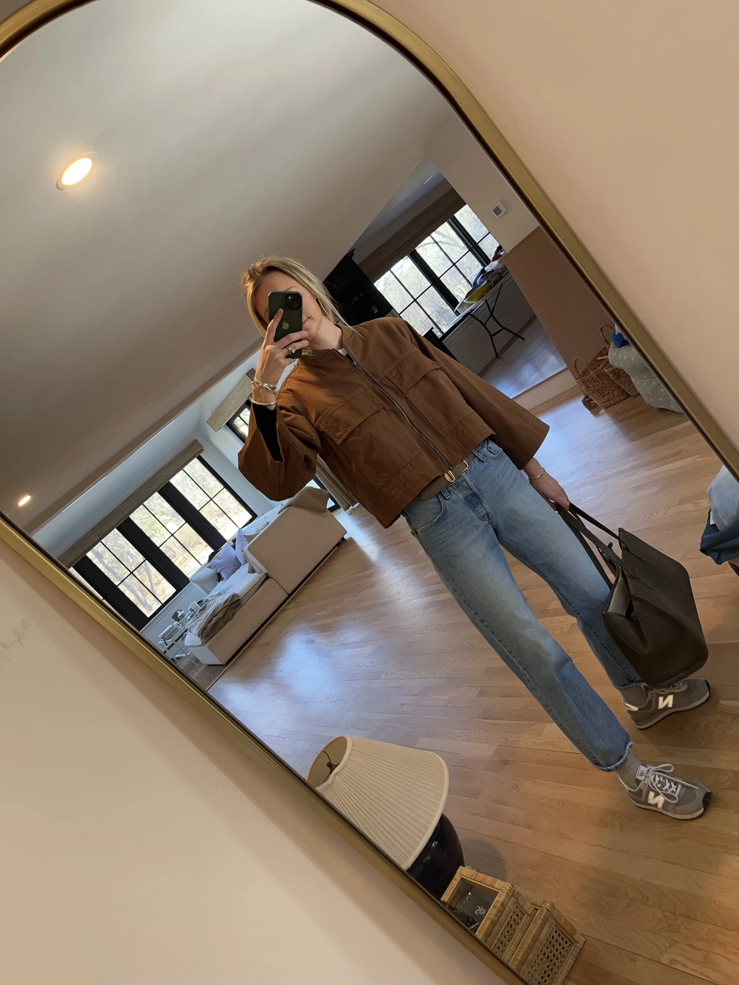 Person taking a mirror selfie in a room with wooden floors, wearing a striped brown and white long-sleeve shirt, light blue jeans, and black and white sneakers.