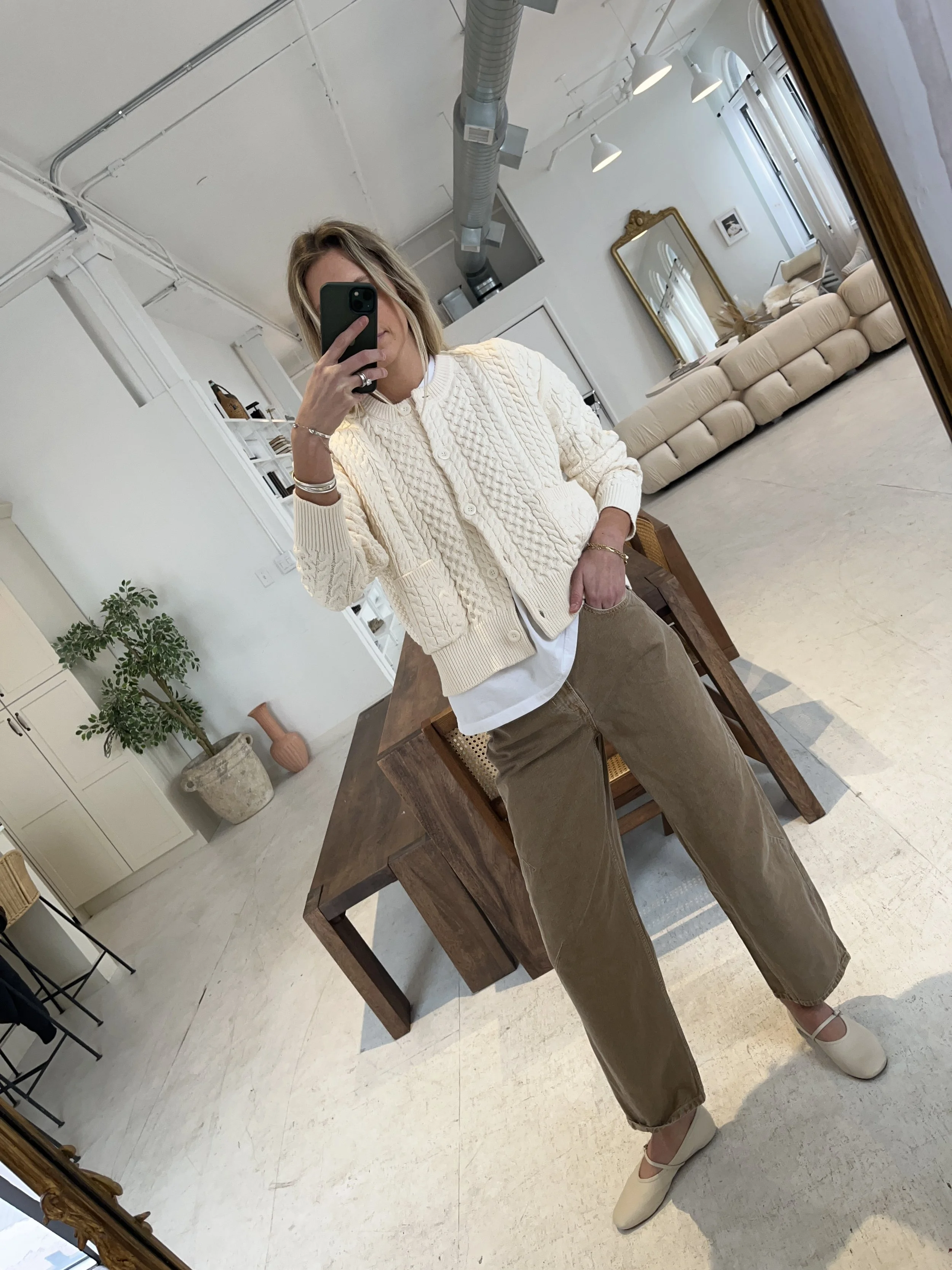 Person taking a mirror selfie in a room with wooden floors, wearing a striped brown and white long-sleeve shirt, light blue jeans, and black and white sneakers.
