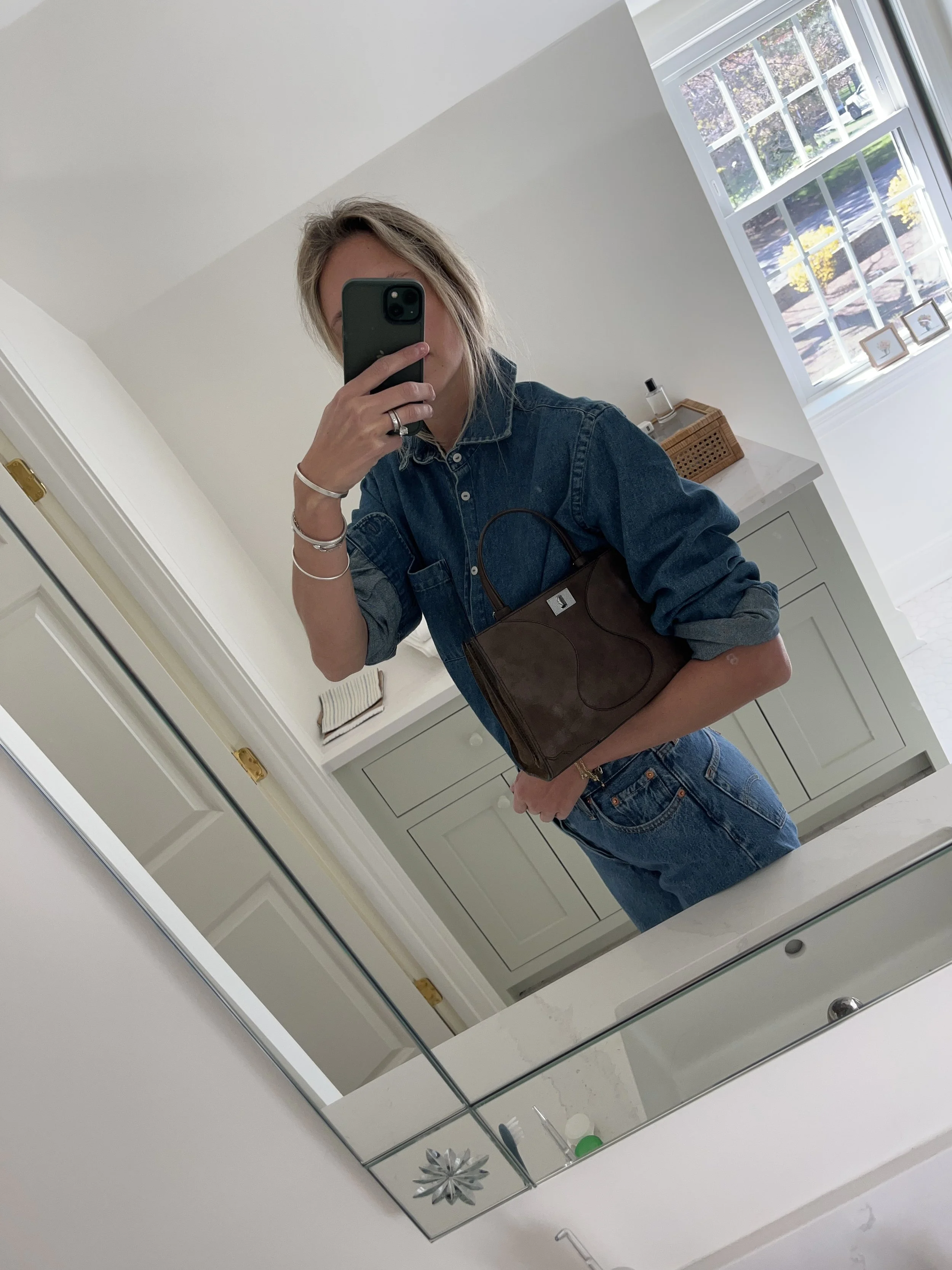 A woman taking a mirror selfie in a spacious living room with wooden floors, large windows, and minimal furniture, wearing a beige jacket, black top, blue jeans, and tan shoes.