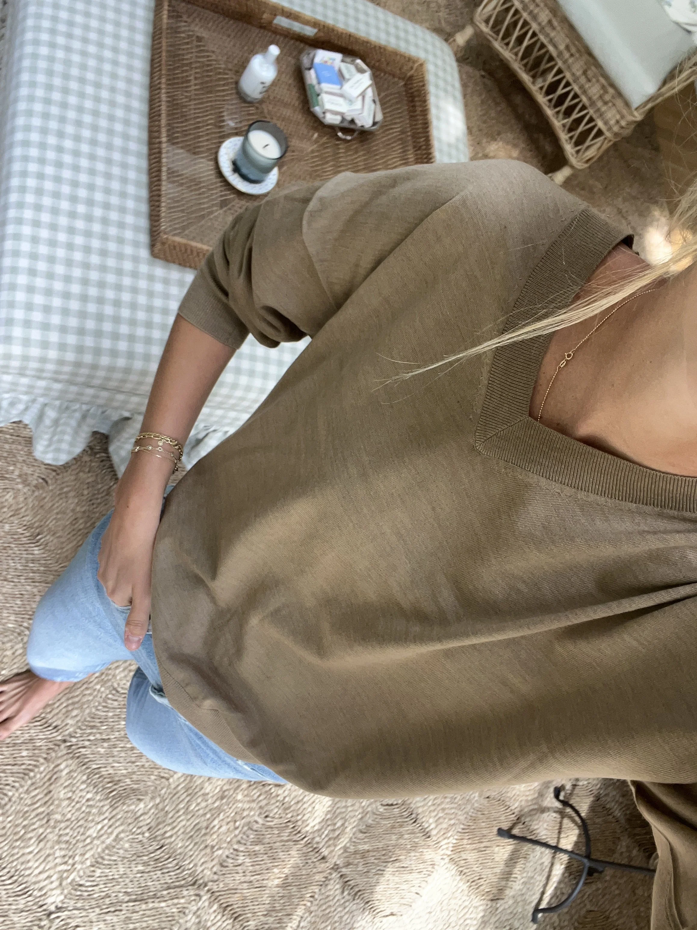 A woman taking a mirror selfie in a spacious living room with wooden floors, large windows, and minimal furniture, wearing a beige jacket, black top, blue jeans, and tan shoes.