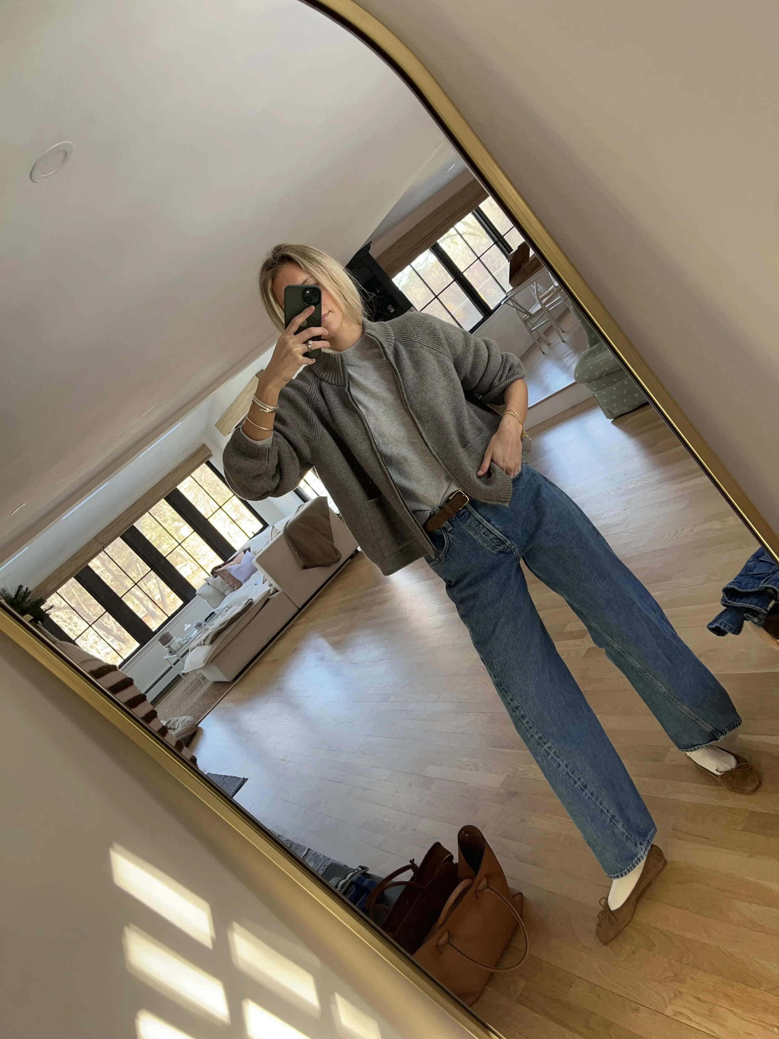Person taking a mirror selfie in a living room with hardwood floors, wearing a black fleece jacket, gray shirt, black leggings, gray sneakers, and a white cap with a logo.