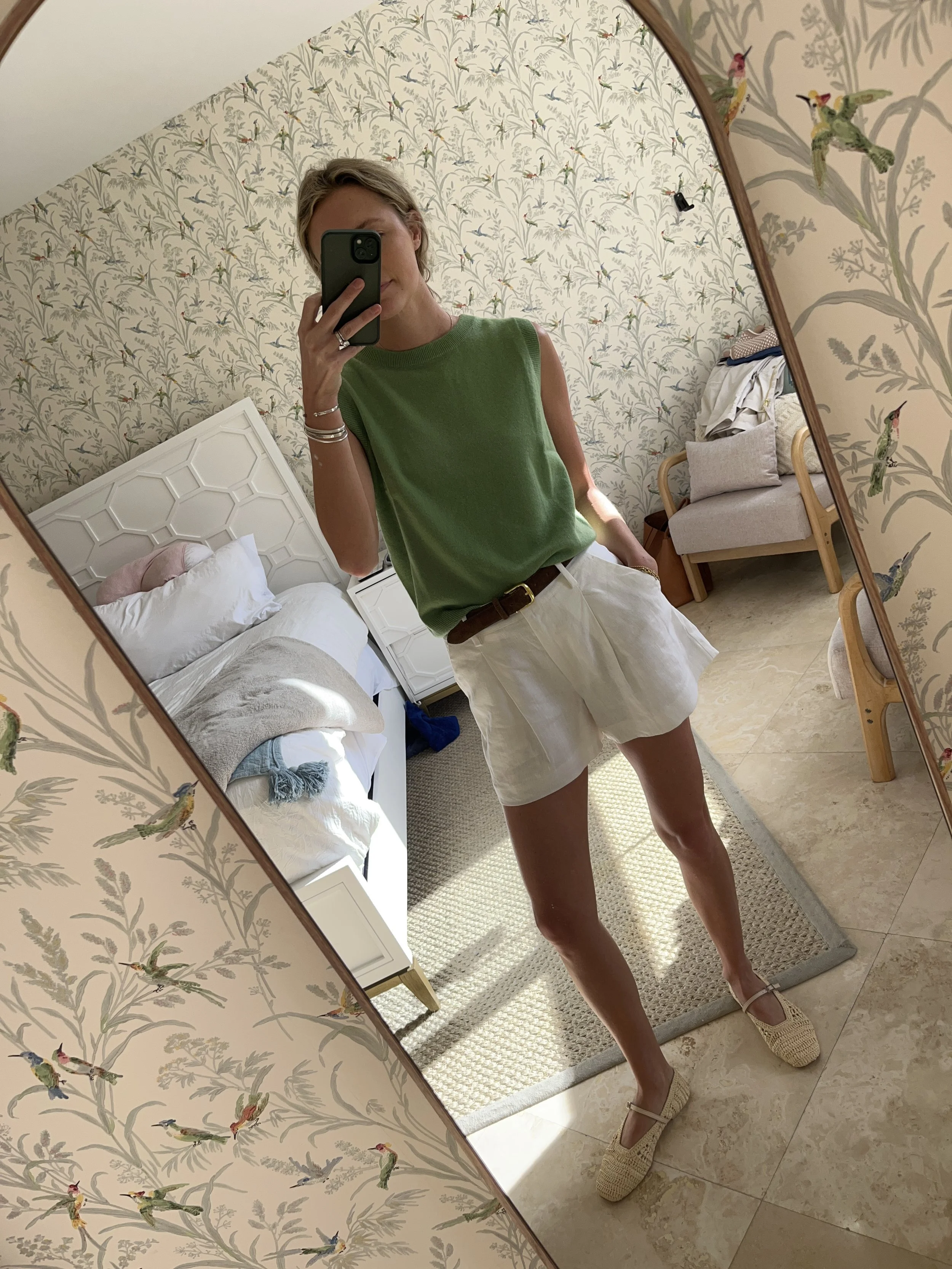 A woman taking a mirror selfie in a spacious living room with wooden floors, large windows, and minimal furniture, wearing a beige jacket, black top, blue jeans, and tan shoes.
