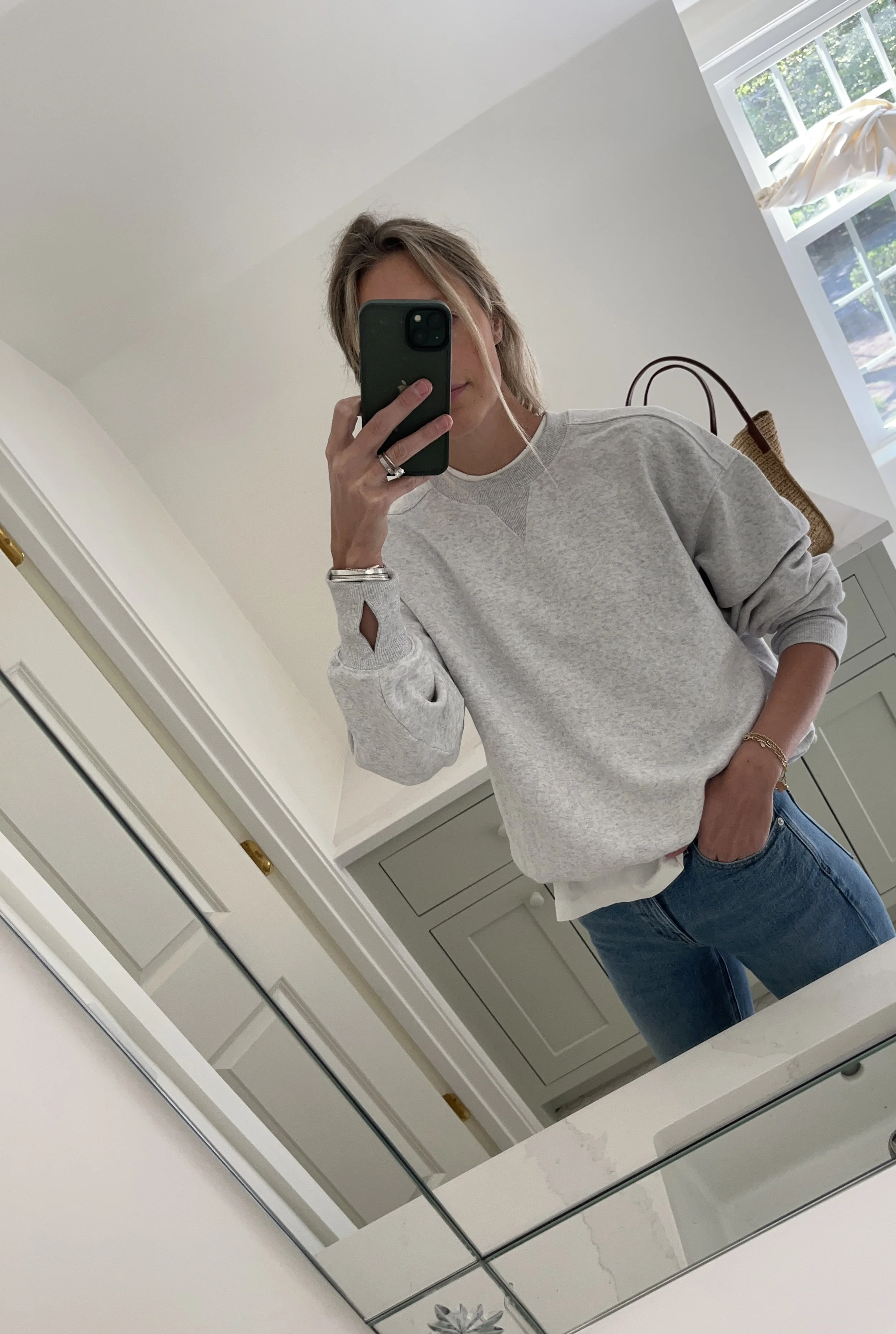 Person taking a mirror selfie in a living room with hardwood floors, wearing a black fleece jacket, gray shirt, black leggings, gray sneakers, and a white cap with a logo.