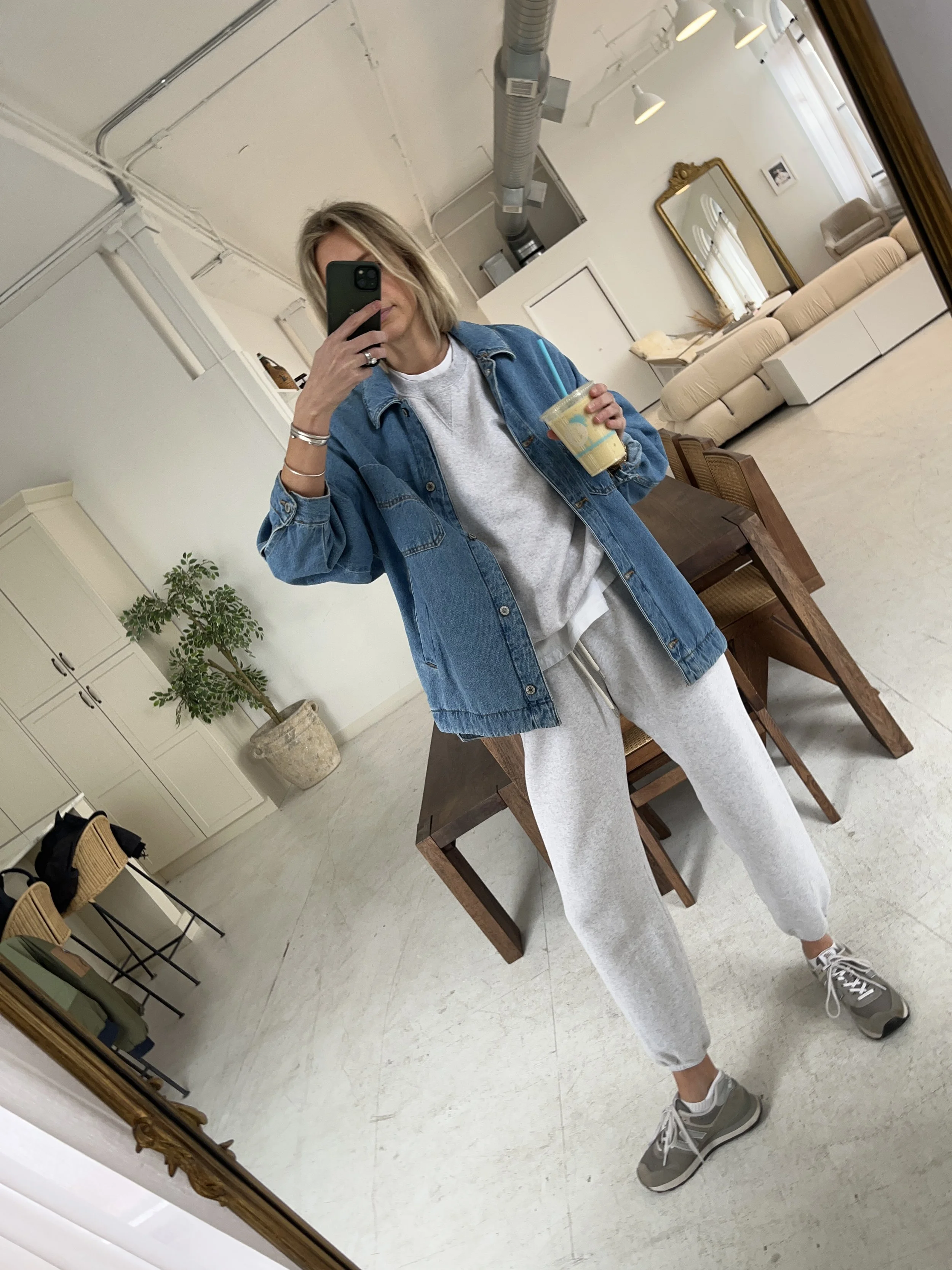 A woman taking a mirror selfie in a spacious living room with wooden floors, large windows, and minimal furniture, wearing a beige jacket, black top, blue jeans, and tan shoes.