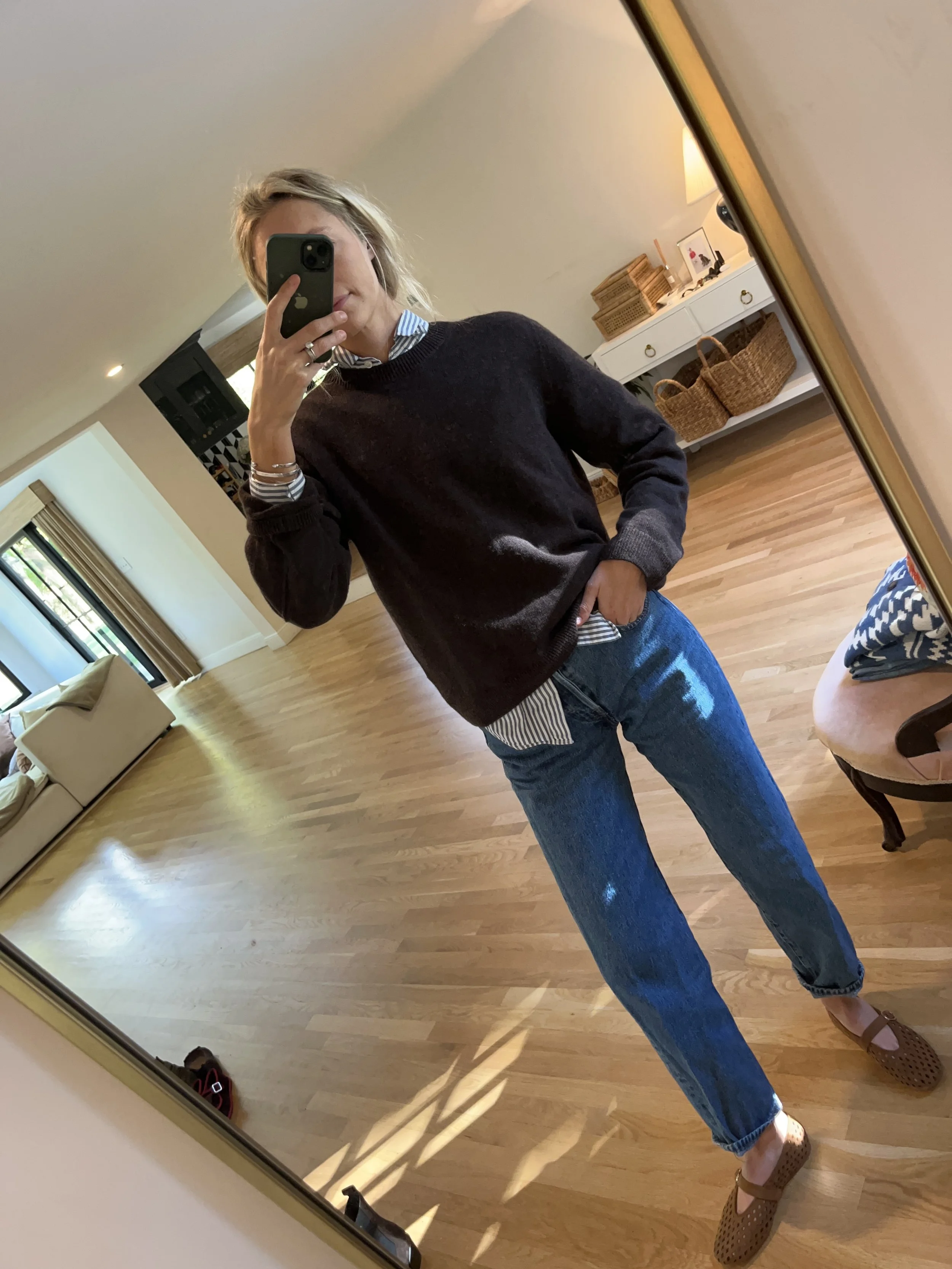 Person taking a mirror selfie in a spacious room with wooden floors, dressed in a dark sweater, striped shirt, blue jeans, and brown shoes.