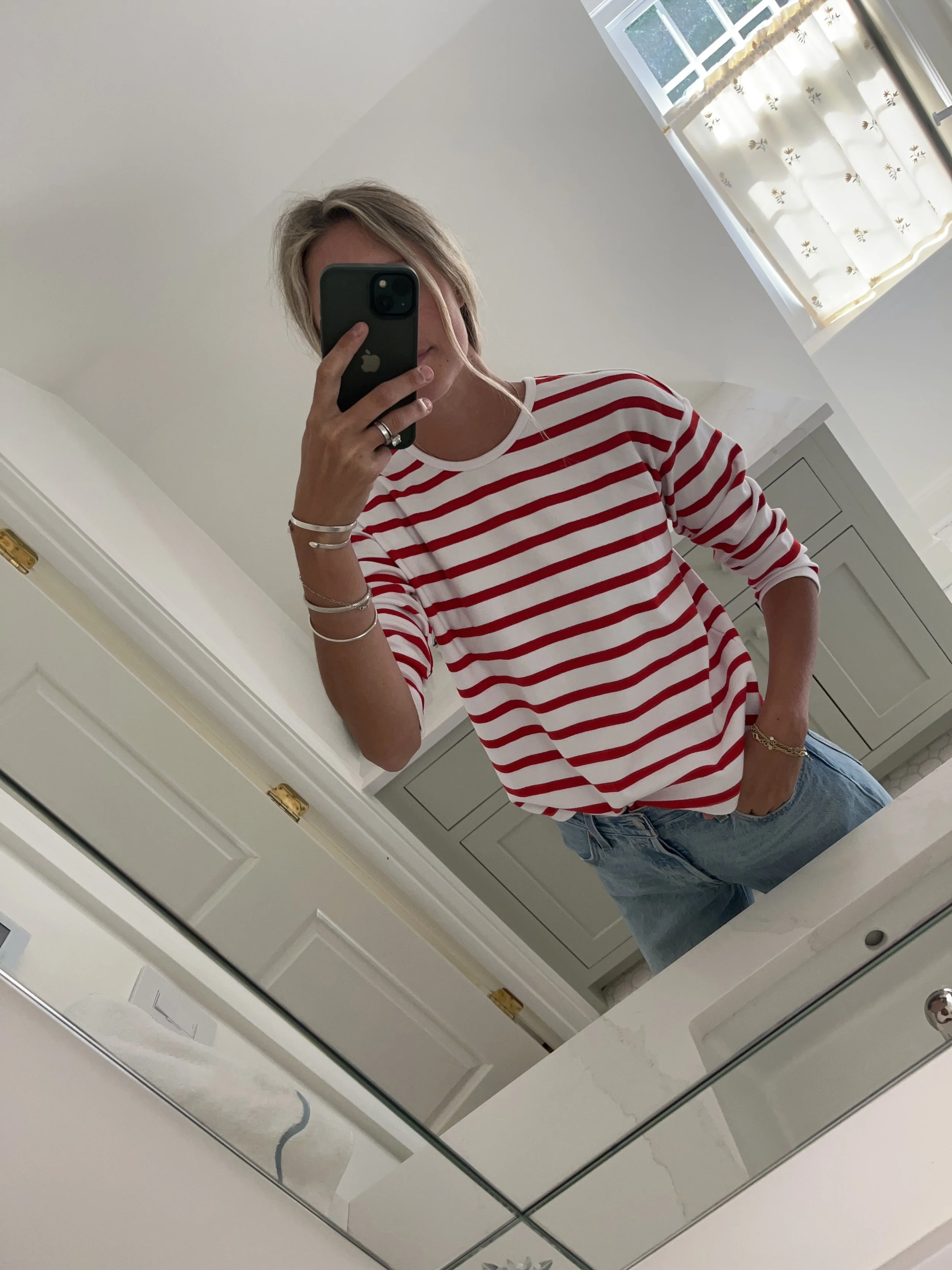 A woman taking a mirror selfie in a spacious living room with wooden floors, large windows, and minimal furniture, wearing a beige jacket, black top, blue jeans, and tan shoes.