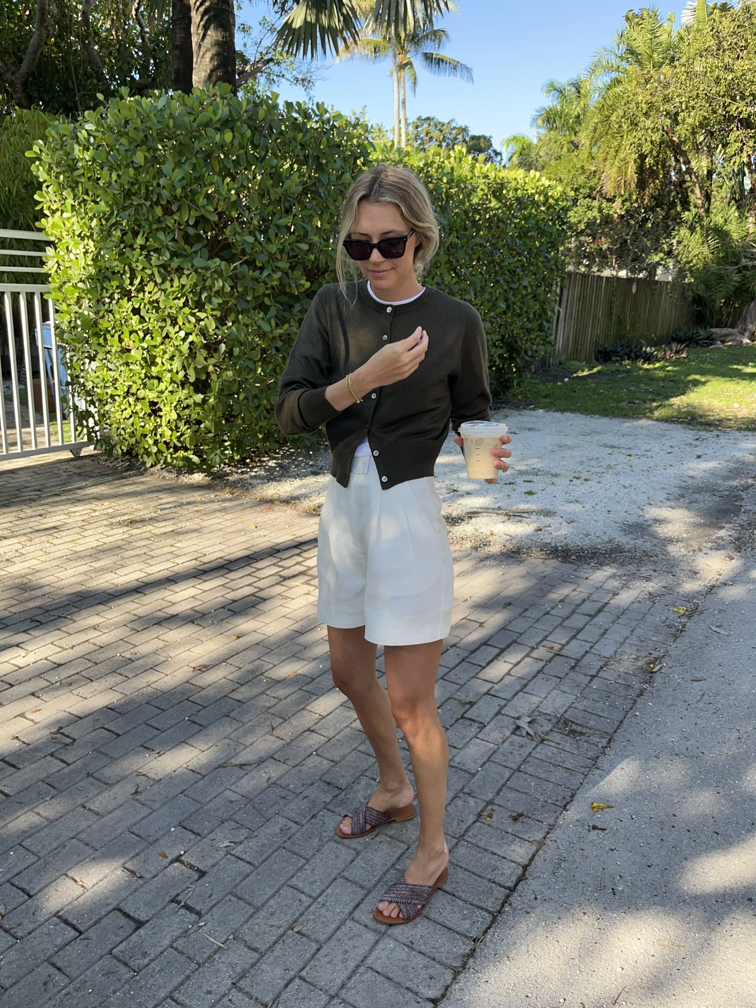 A woman taking a mirror selfie in a spacious living room with wooden floors, large windows, and minimal furniture, wearing a beige jacket, black top, blue jeans, and tan shoes.