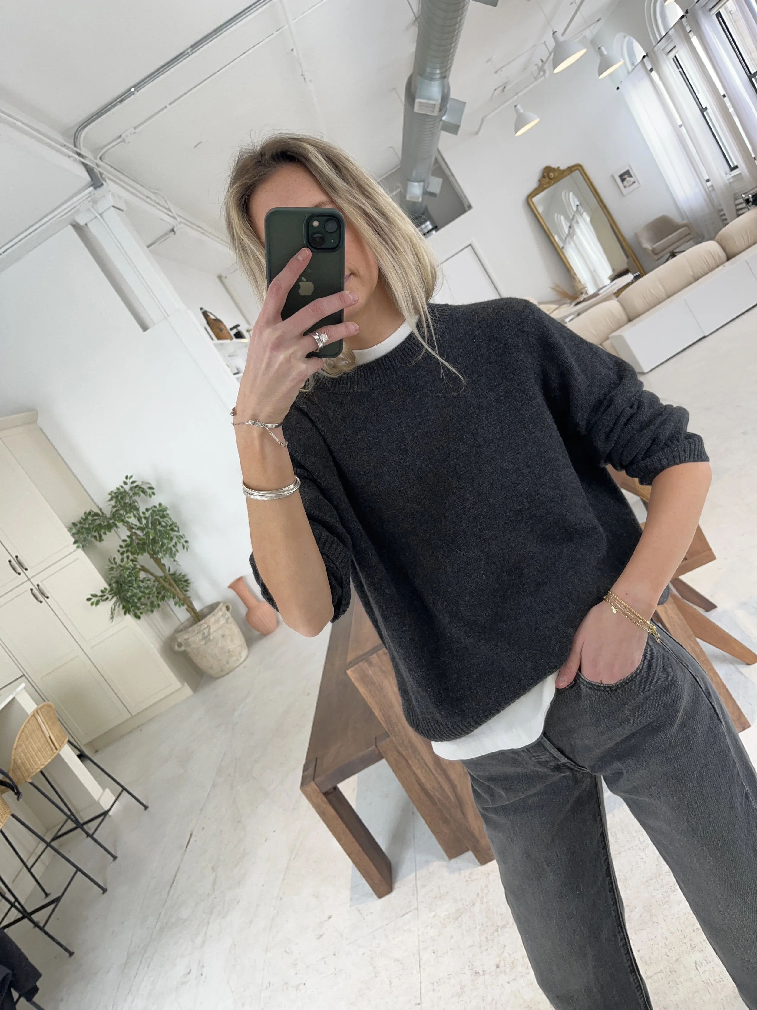 Person taking a mirror selfie in a living room with hardwood floors, wearing a black fleece jacket, gray shirt, black leggings, gray sneakers, and a white cap with a logo.