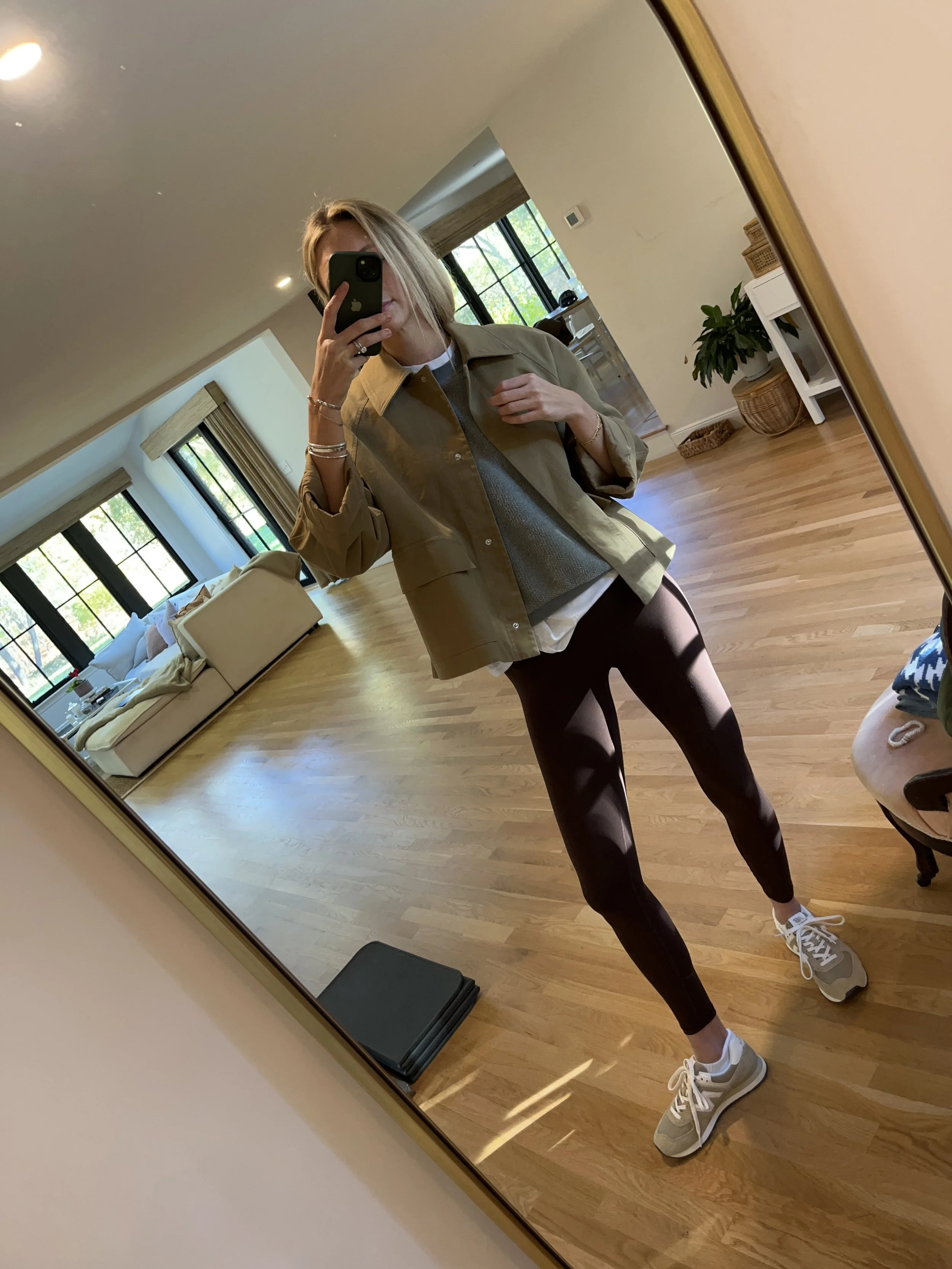 A woman taking a mirror selfie in a well-lit living room, wearing a beige jacket, gray sweater, black leggings, and gray sneakers.