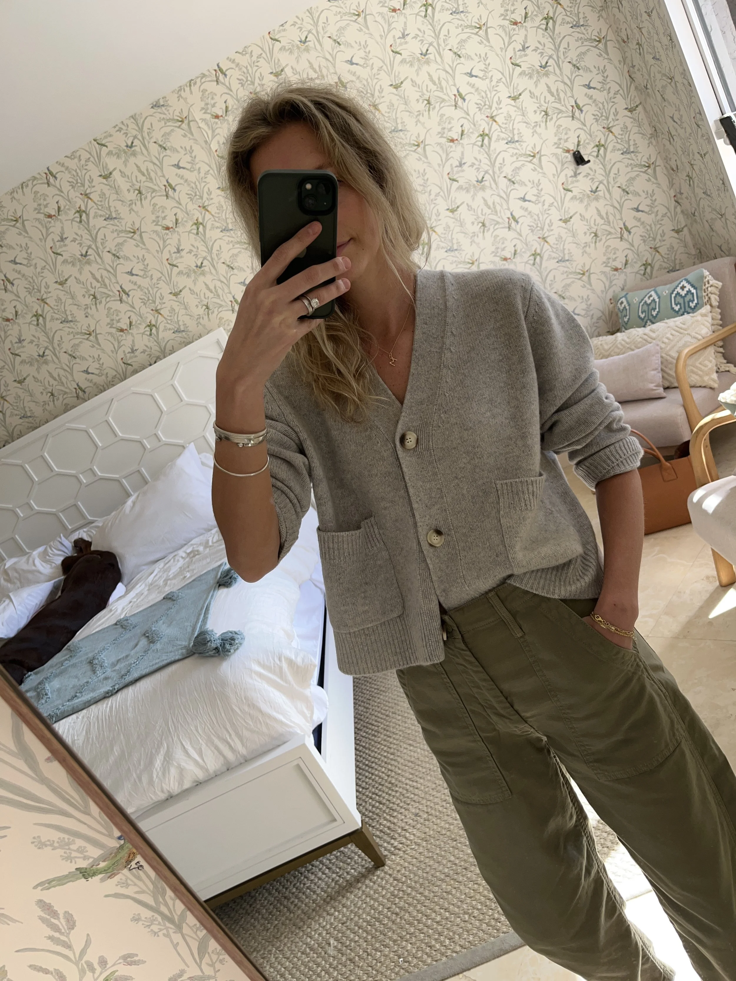 A woman taking a mirror selfie in a spacious, well-lit living room with hardwood floors, large windows, a white sofa, and a side table with a plant.