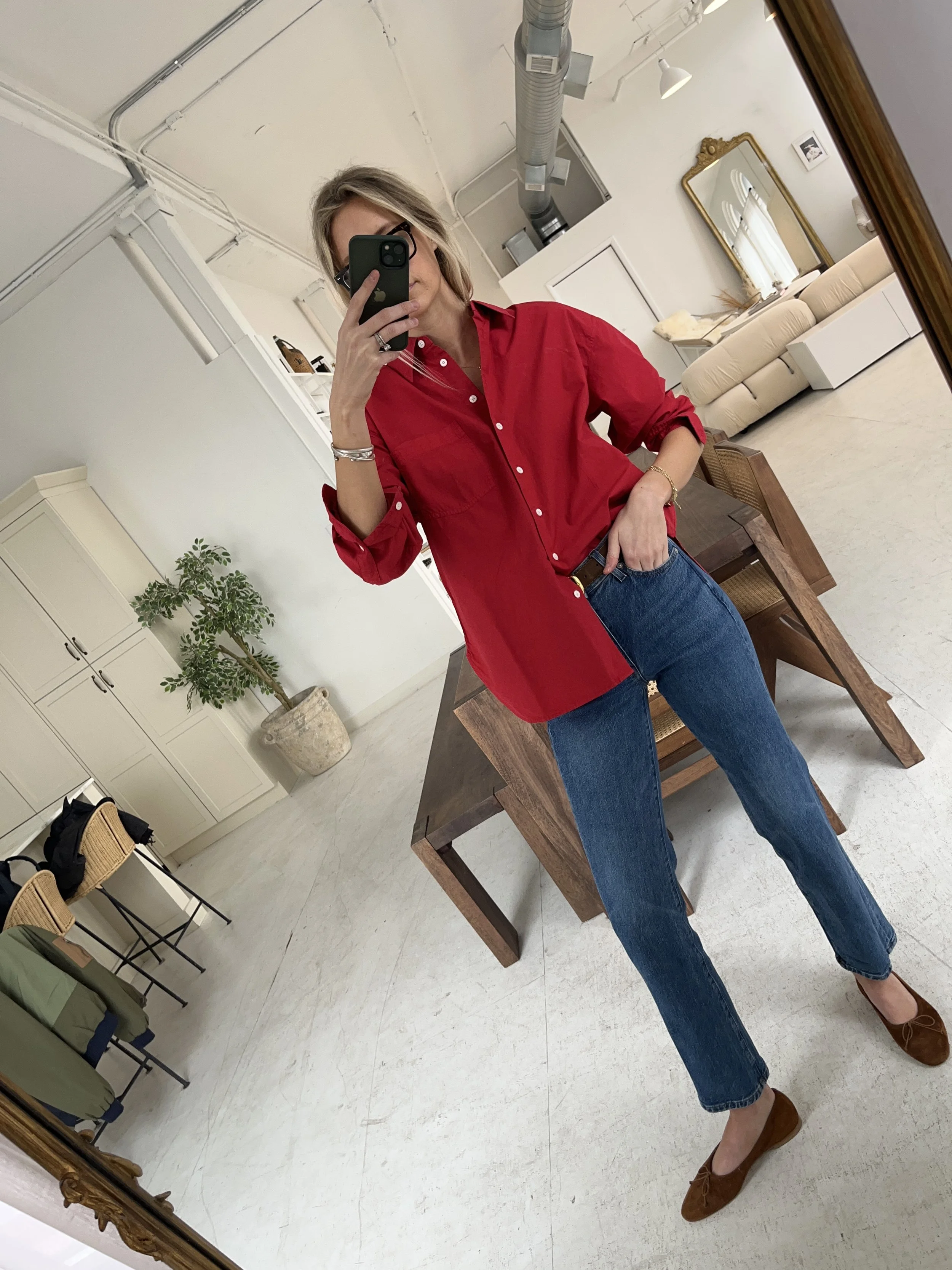 Person taking a mirror selfie in a living room with hardwood floors, wearing a black fleece jacket, gray shirt, black leggings, gray sneakers, and a white cap with a logo.