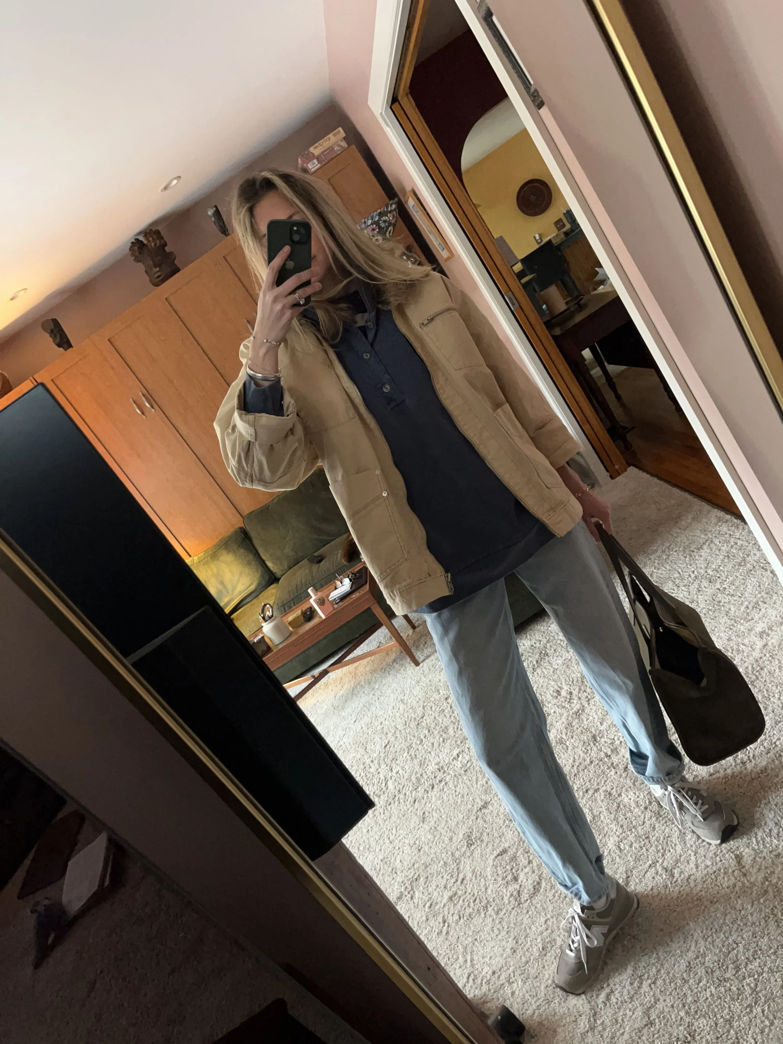 A woman taking a mirror selfie in a spacious, well-lit living room with hardwood floors, large windows, a white sofa, and a side table with a plant.