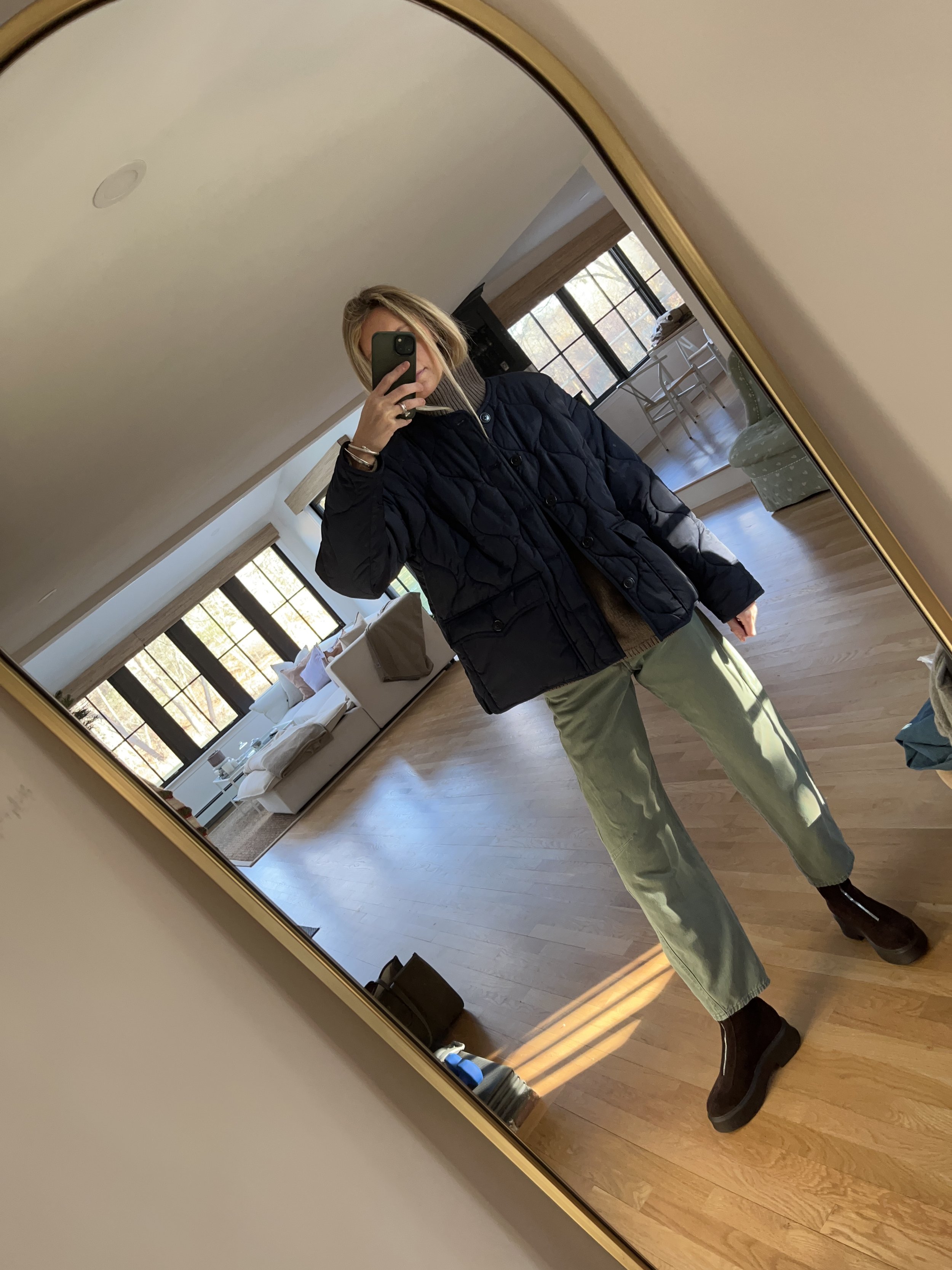A woman taking a mirror selfie in a spacious living room with wooden floors, large windows, and minimal furniture, wearing a beige jacket, black top, blue jeans, and tan shoes.