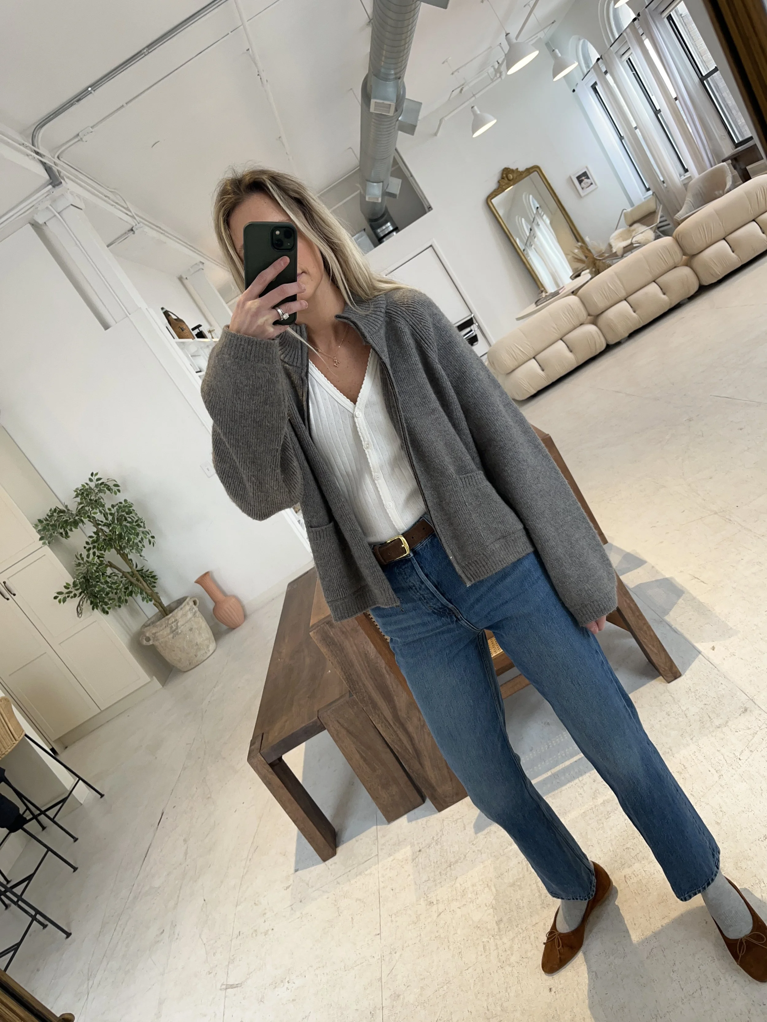 A woman taking a mirror selfie in a spacious, well-lit living room with hardwood floors, large windows, a white sofa, and a side table with a plant.