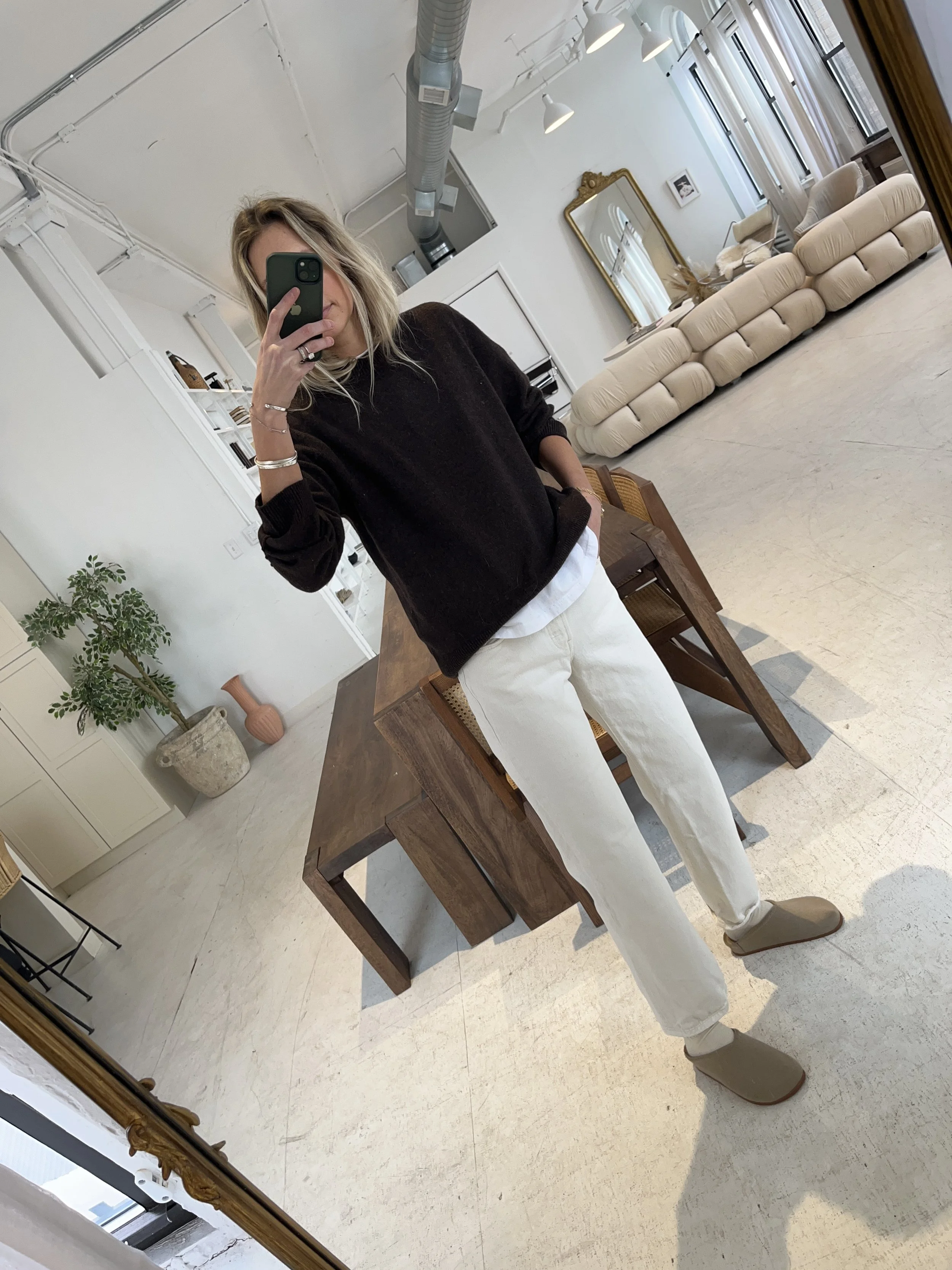 Person taking a mirror selfie in a living room with hardwood floors, wearing a black fleece jacket, gray shirt, black leggings, gray sneakers, and a white cap with a logo.