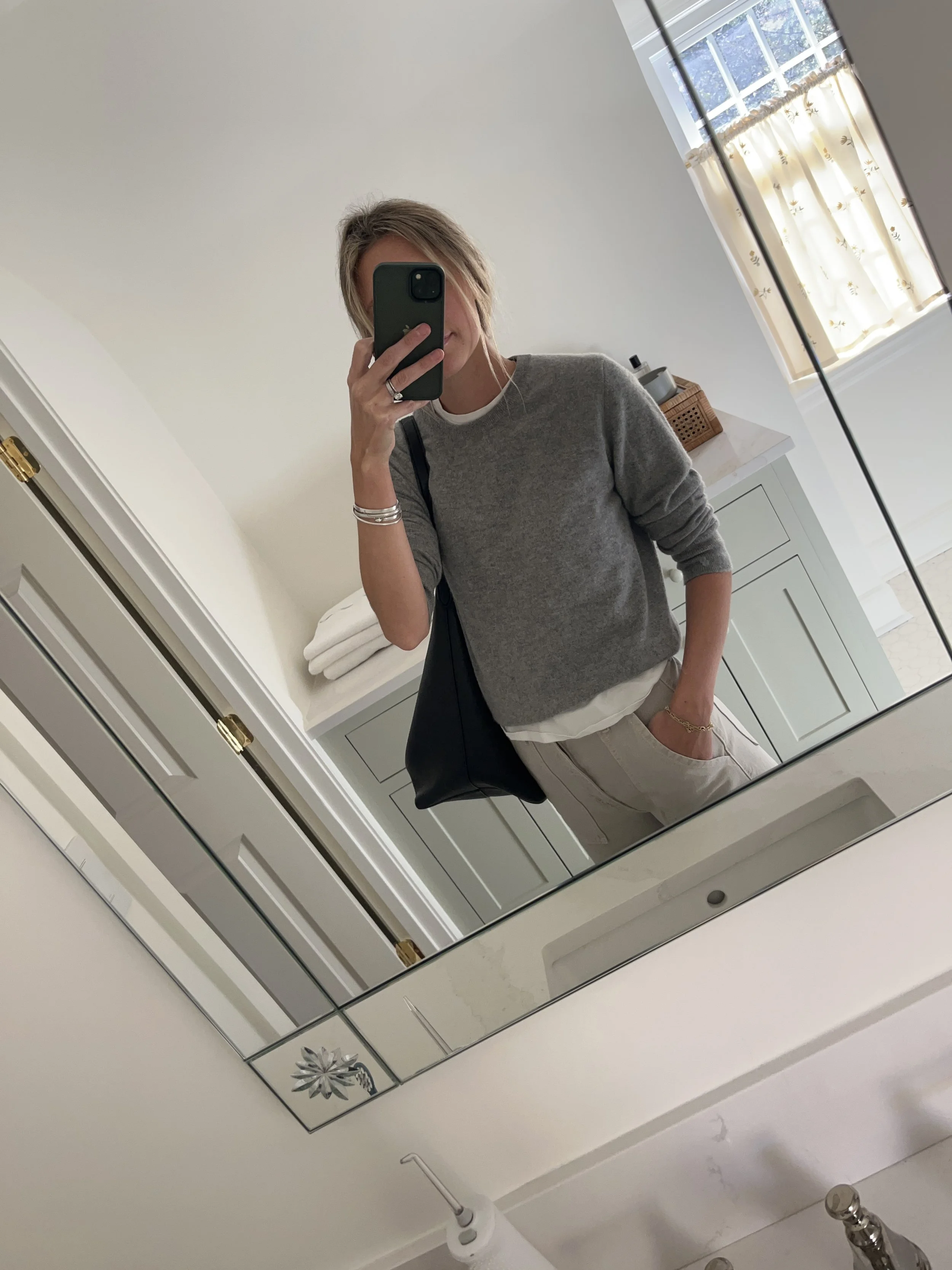 A woman taking a mirror selfie in a spacious, well-lit living room with hardwood floors, large windows, a white sofa, and a side table with a plant.