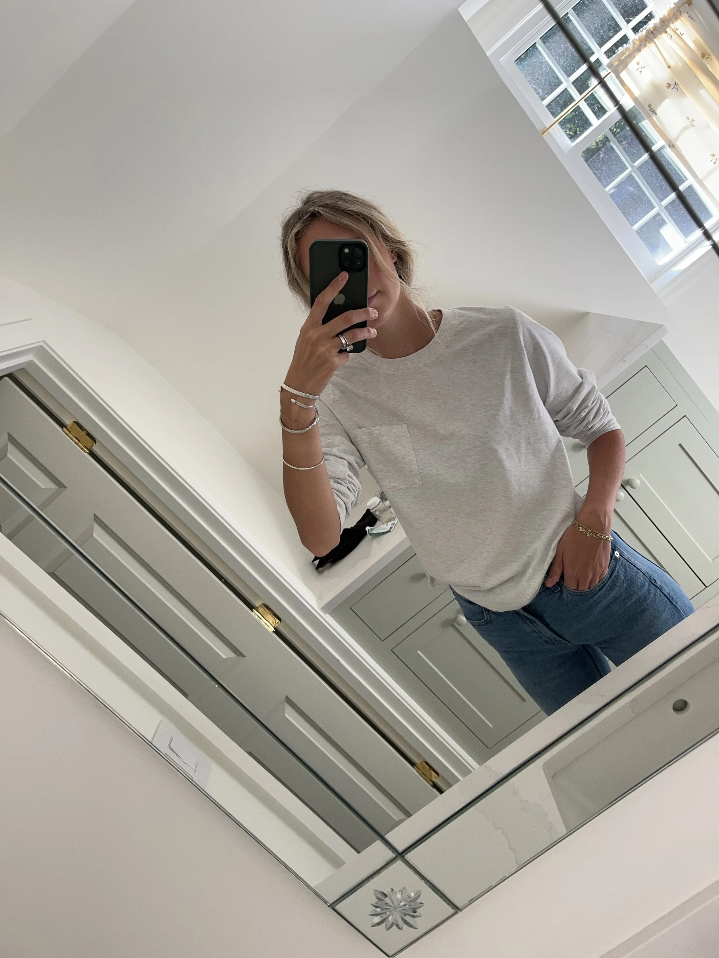 A woman taking a mirror selfie in a spacious living room with wooden floors, large windows, and minimal furniture, wearing a beige jacket, black top, blue jeans, and tan shoes.