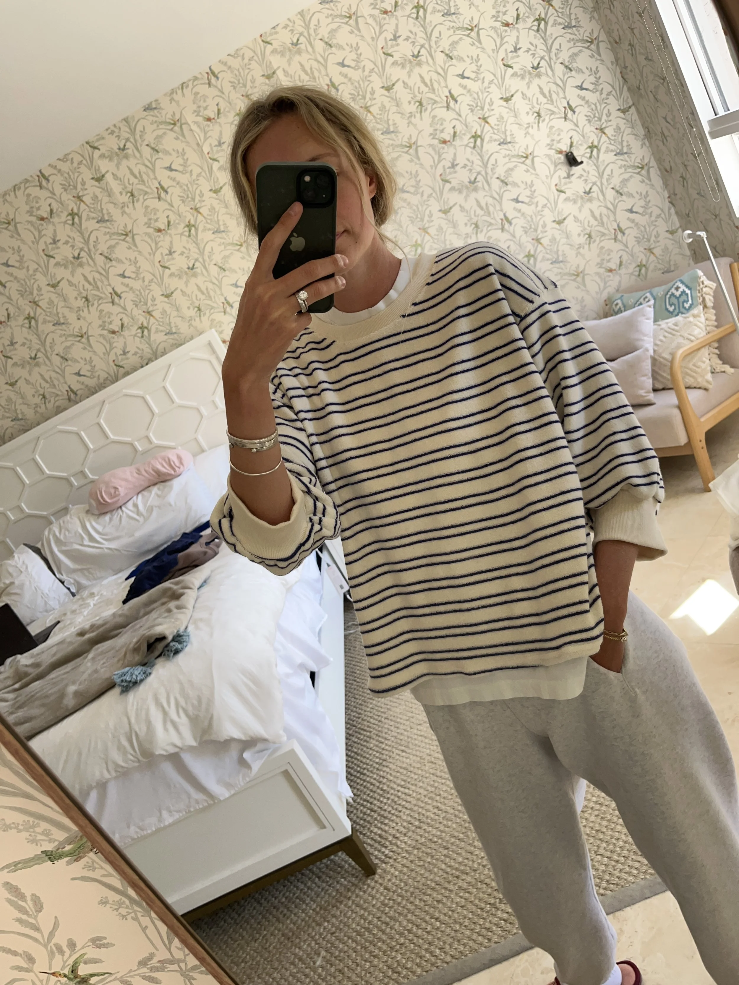 Person taking a mirror selfie in a living room with hardwood floors, wearing a black fleece jacket, gray shirt, black leggings, gray sneakers, and a white cap with a logo.