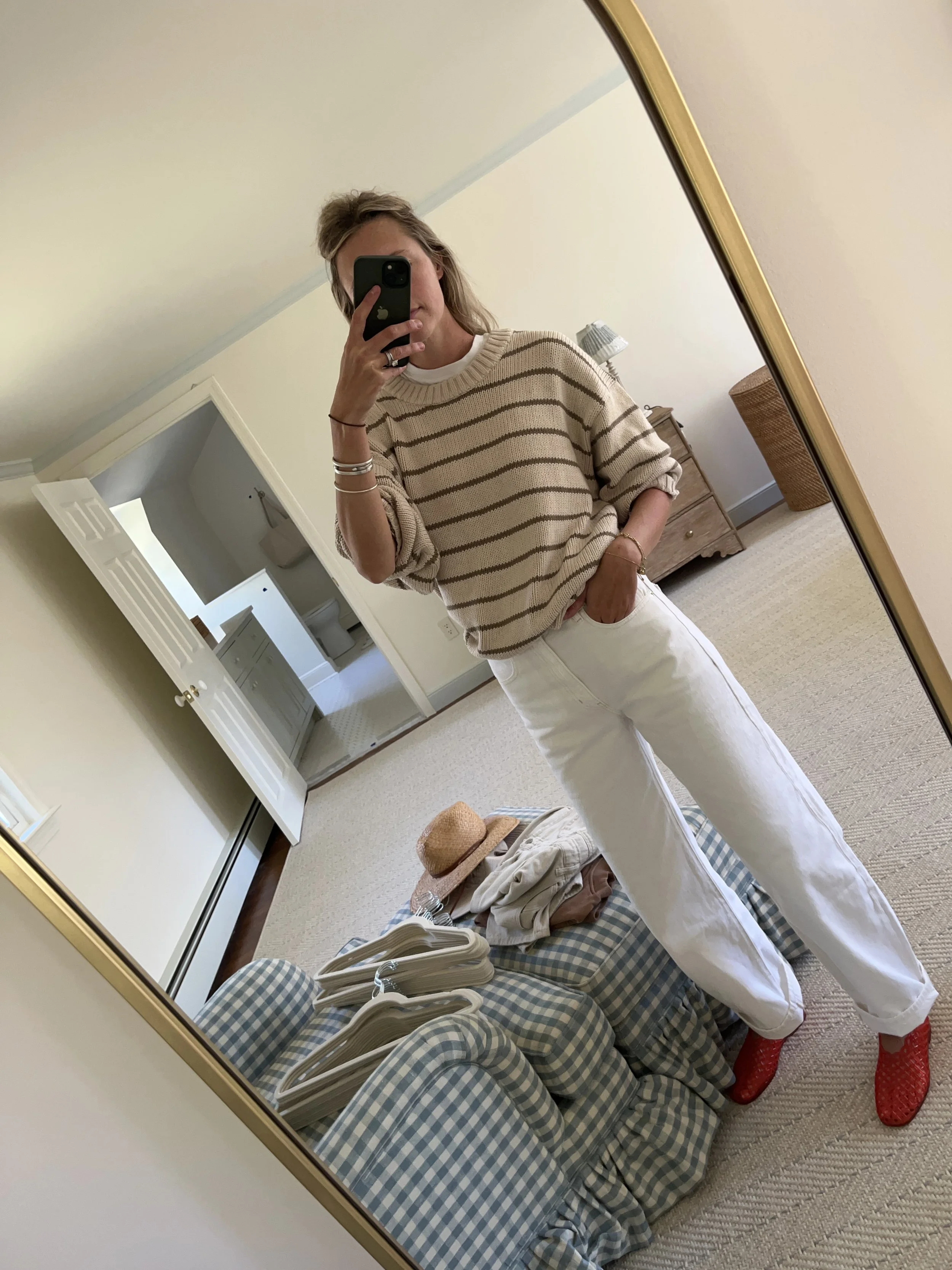 Person taking a mirror selfie in a living room with hardwood floors, wearing a black fleece jacket, gray shirt, black leggings, gray sneakers, and a white cap with a logo.