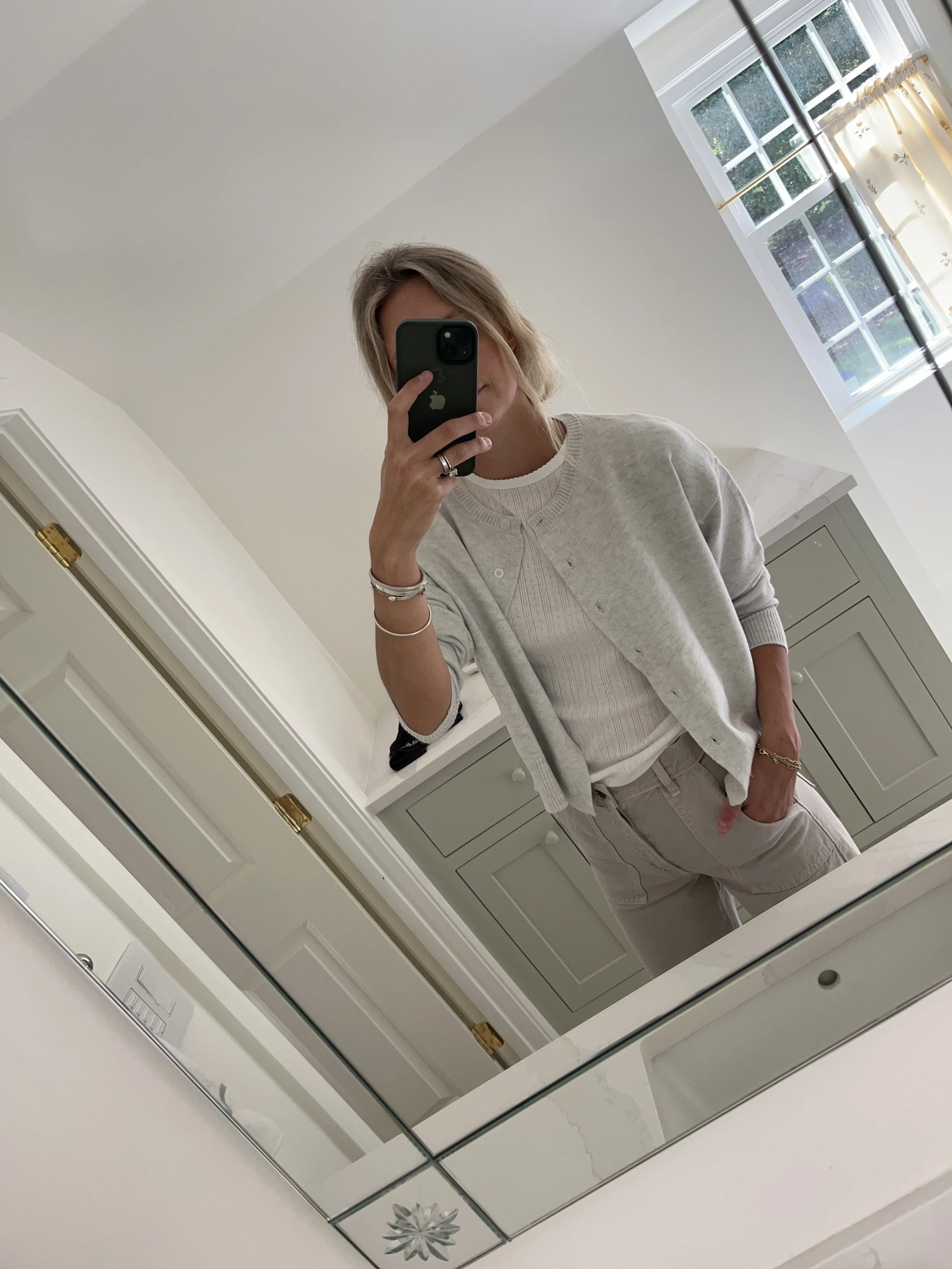 A woman taking a mirror selfie in a spacious living room with wooden floors, large windows, and minimal furniture, wearing a beige jacket, black top, blue jeans, and tan shoes.