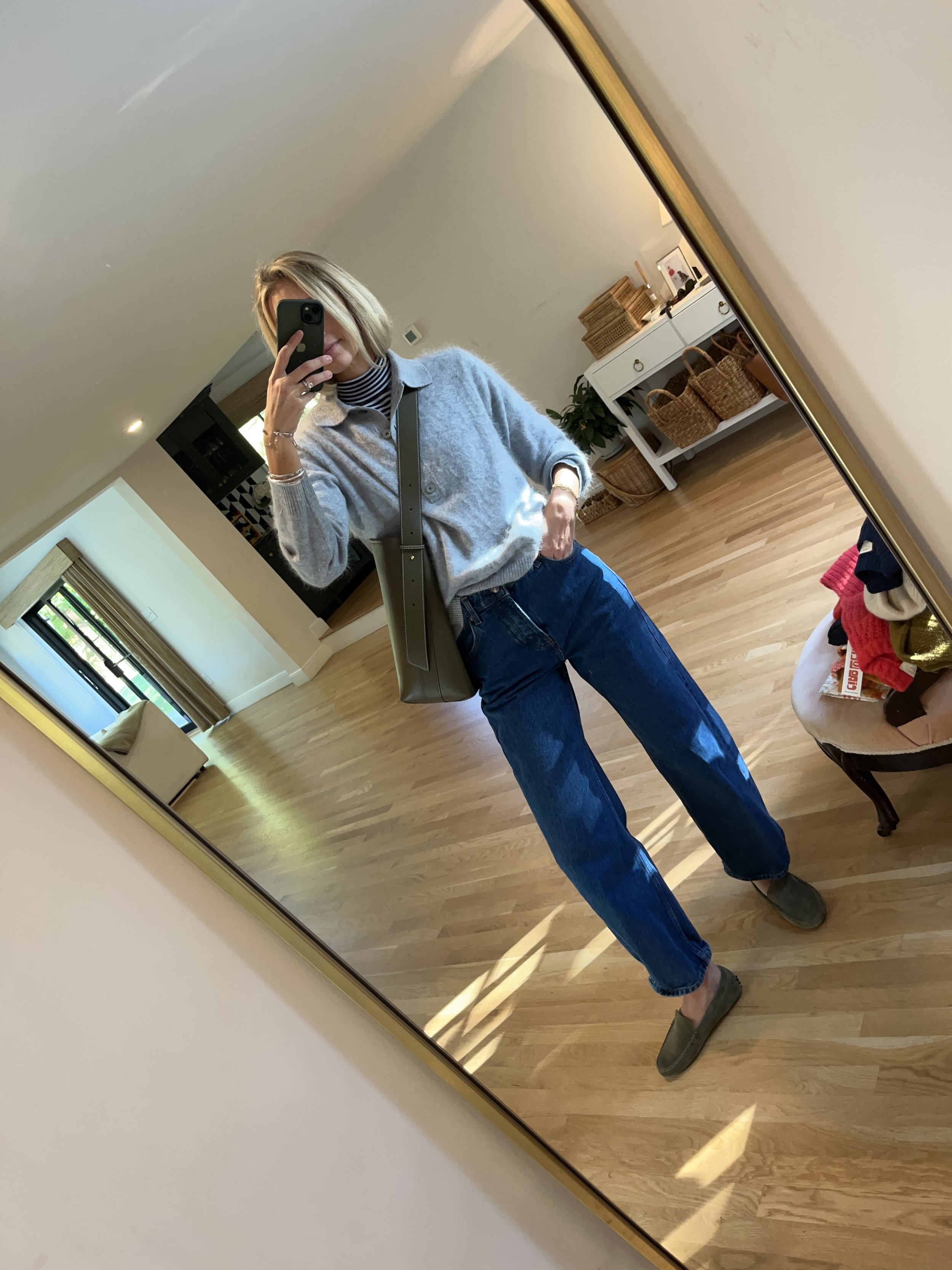 A woman taking a mirror selfie in a spacious, well-lit living room with wooden floors, wearing a gray sweater over a striped top, blue jeans, and green shoes, with a gray crossbody bag.