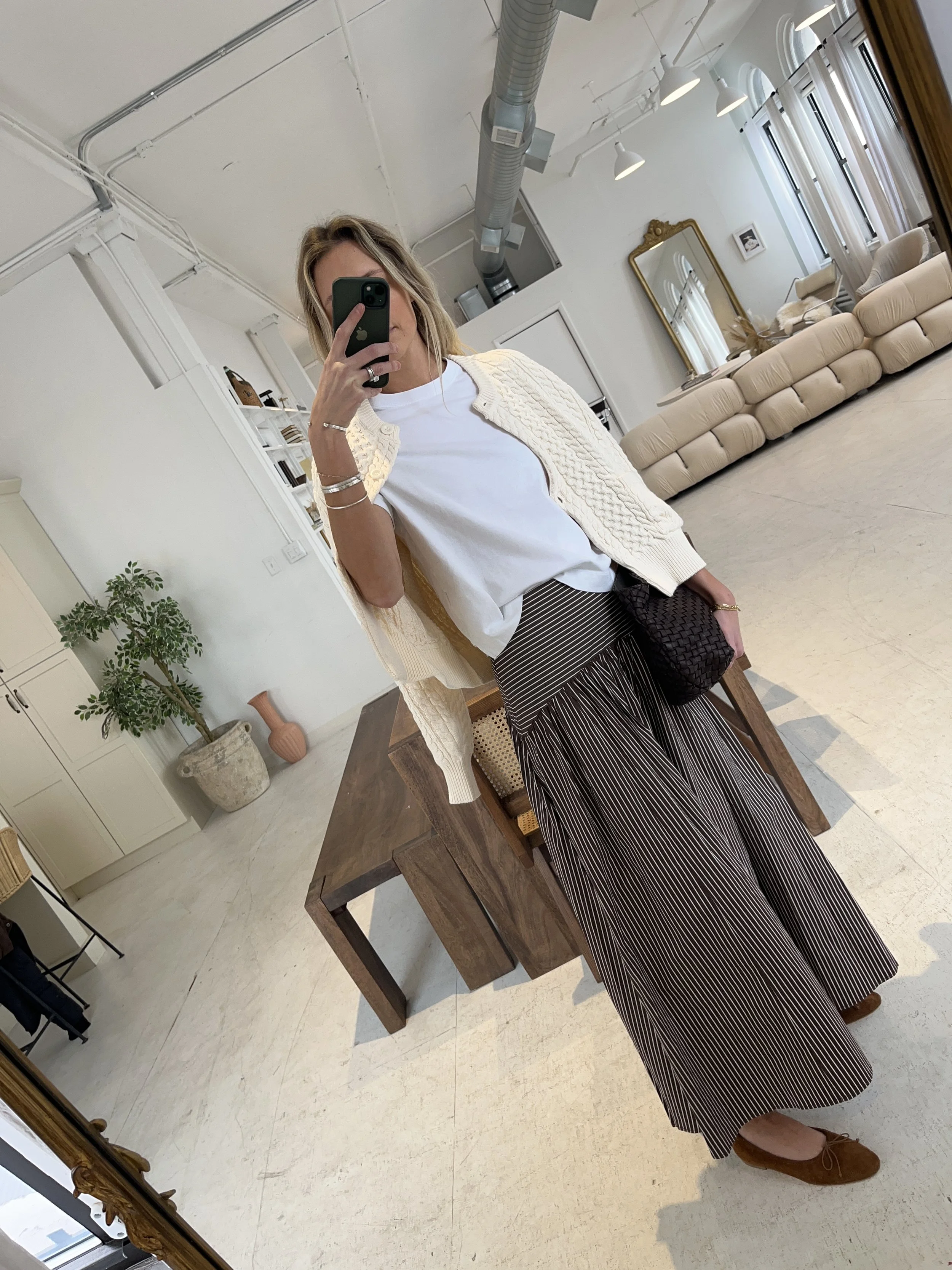 A woman taking a mirror selfie in a spacious, well-lit living room with hardwood floors, large windows, a white sofa, and a side table with a plant.